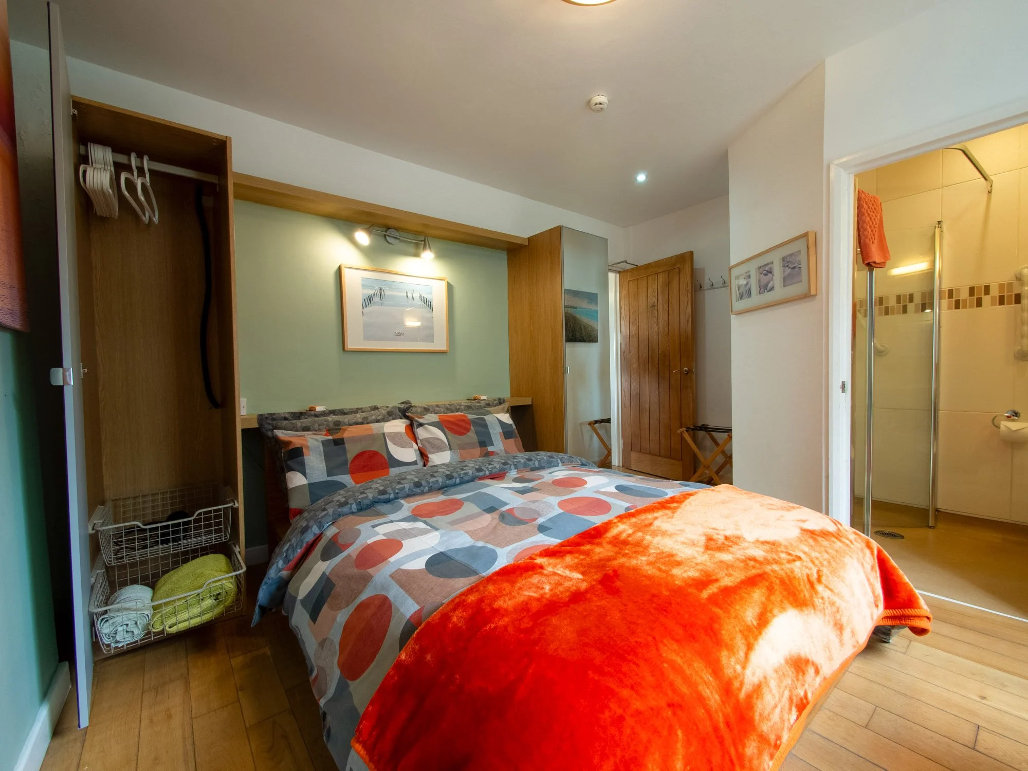 A cozy bedroom with a bed that has a colorful geometric-patterned duvet and orange plush blanket. There is a wooden wardrobe and a small open closet shelf with towels and hangers. The room features framed seaside photographs on the wall and an adjace
