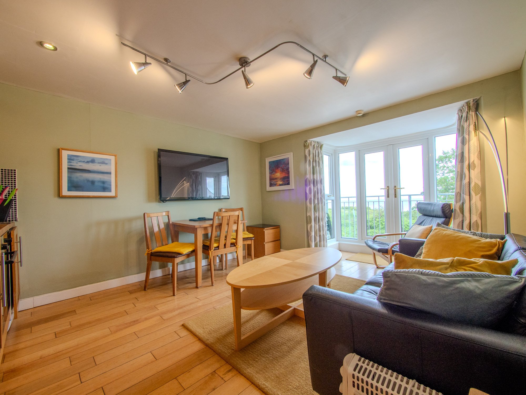 Living room with a sofa, coffee table, dining table with chairs, wall-mounted TV, paintings, large window with curtains, and hardwood floor.