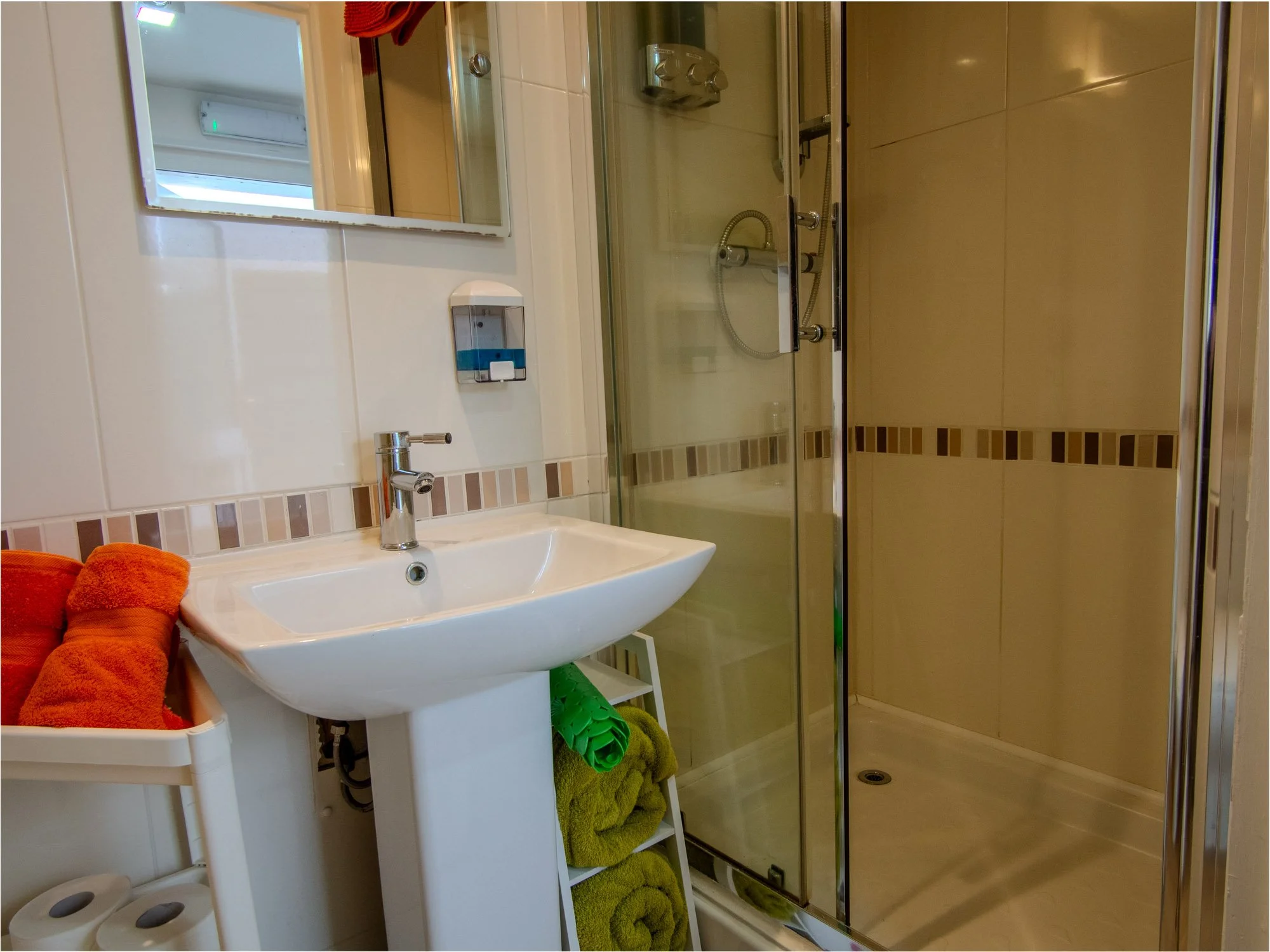 A small bathroom with a sink, mirror, towel rack, and a shower stall. The towel rack holds orange towels, and green towels are stacked beneath the sink. The shower has a glass door and a showerhead, with tiled walls and a drain on the floor.