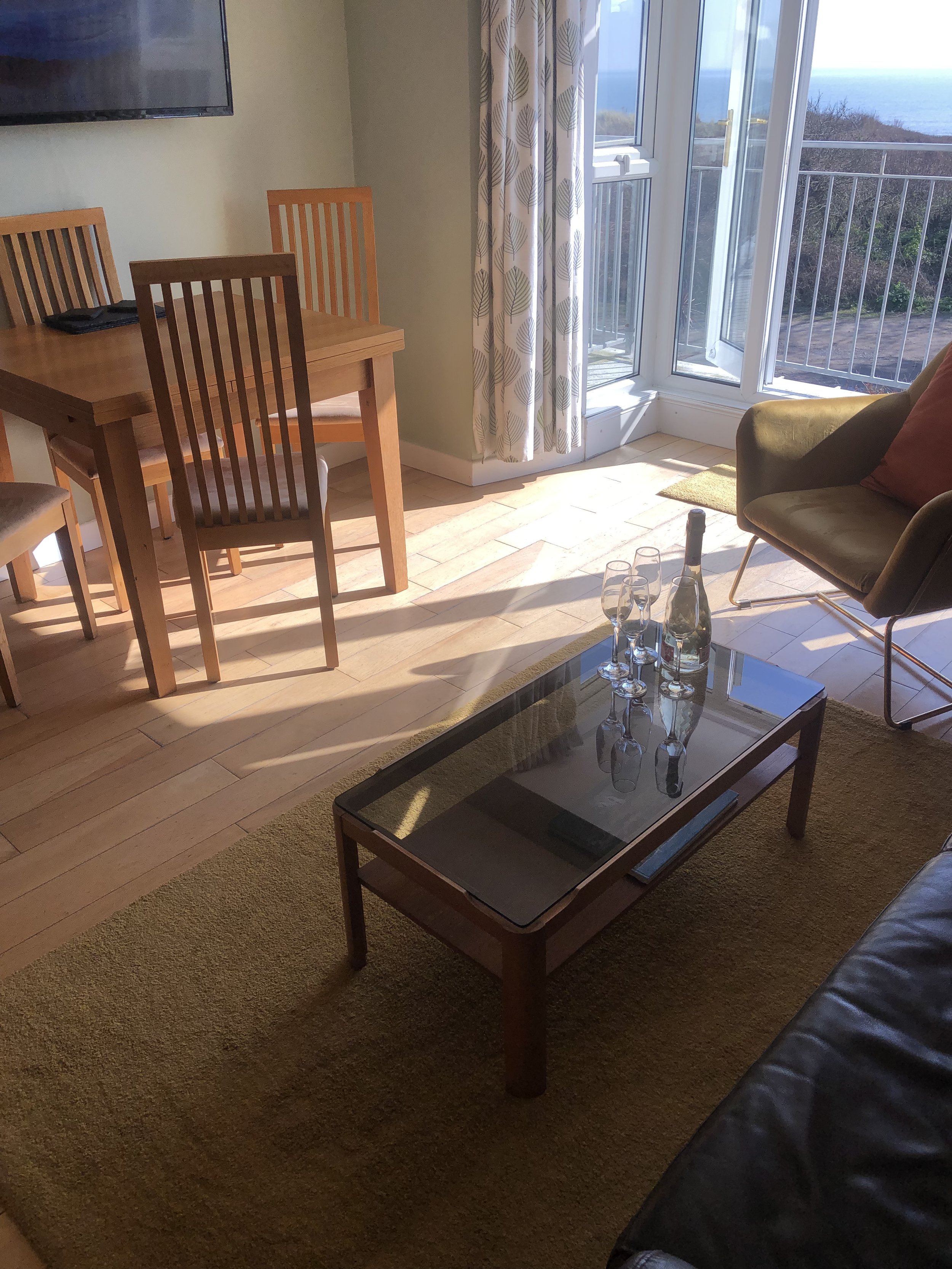 Room with dining table, chairs, and a glass coffee table with wine bottle and glasses; sunny view with patio doors and ocean in background.