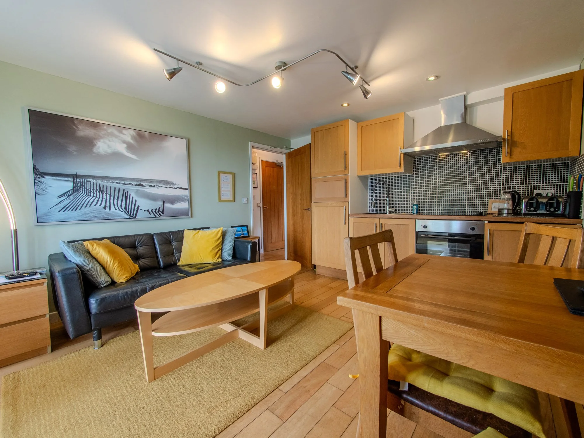 A living room and kitchen area with light green walls, a framed black-and-white beach photograph on the wall, a black leather sofa with yellow and gray cushions, a wooden coffee table, and wooden dining chairs. The kitchen has wooden cabinets, a blac