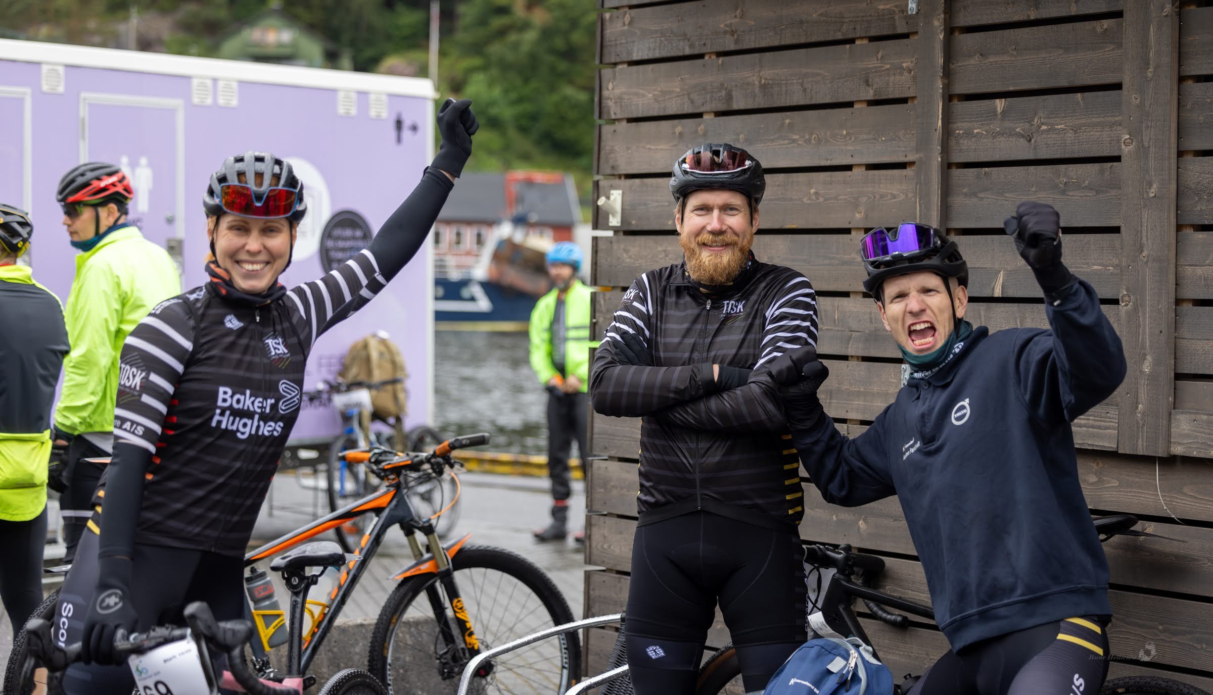 Three cyclists in cycling gear and helmets posing with arms raised and smiling; two men and a woman, with bikes, near a wooden wall and a body of water in the background.
