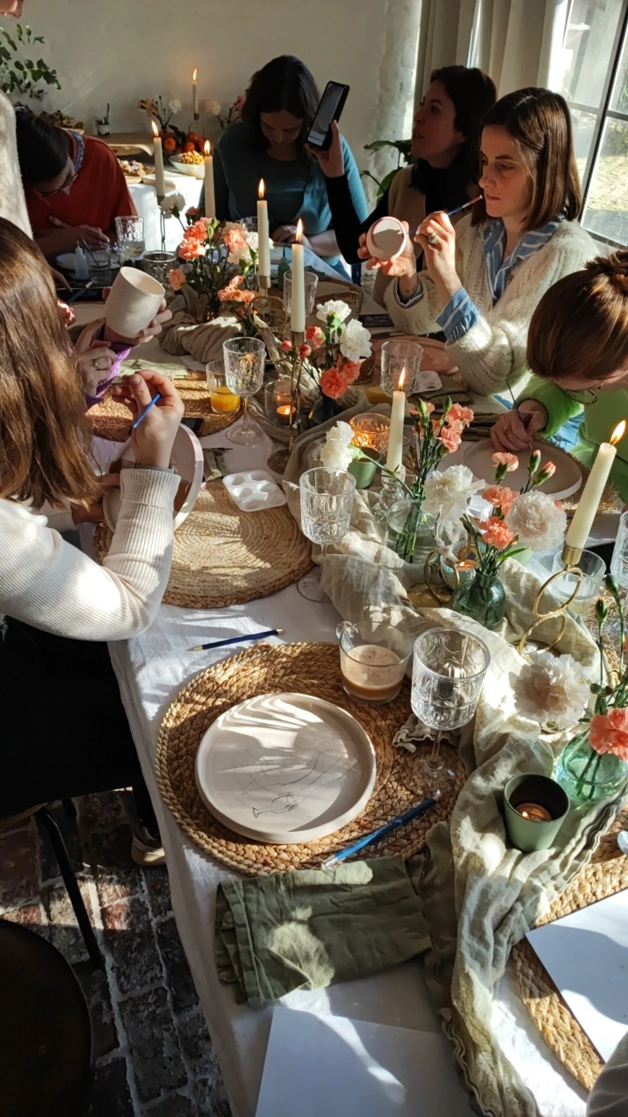 Mensen zitten rond een tafel met bloemen, kaarsen, borden en potjes verf. Ze zijn bezig met schilderen of decoreren. Er is natuurlijk licht en de sfeer is gezellig.