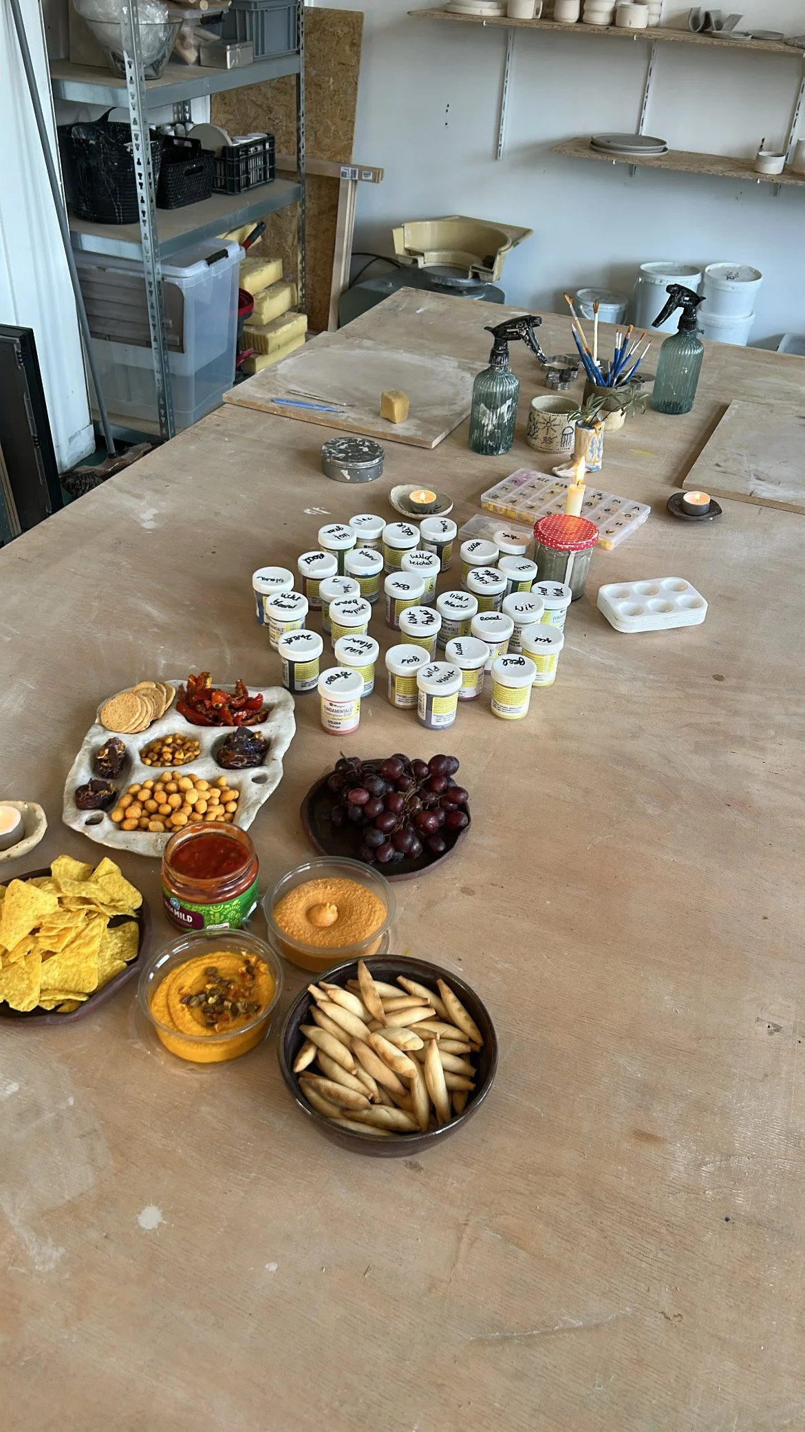 Een werktafel met verschillende potten, borden met snacks, druiven, en kleine containers met voedsel, in een atelier of keukenruimte.