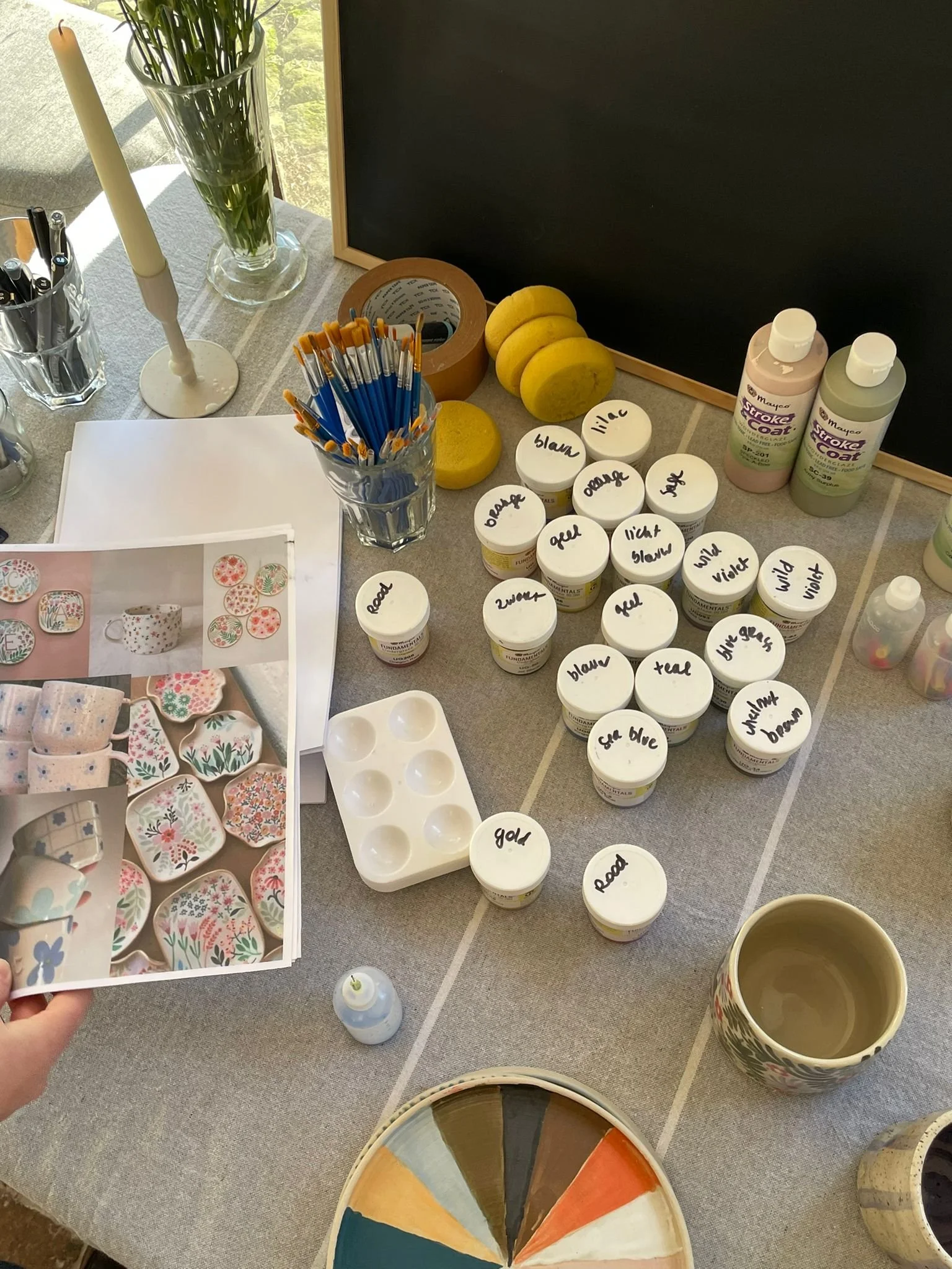 Tafel met schilderbenodigdheden zoals kwasten, verfpotjes met labels, voorbeeldfoto's van beschilderd aardewerk en een kleurenpalet.