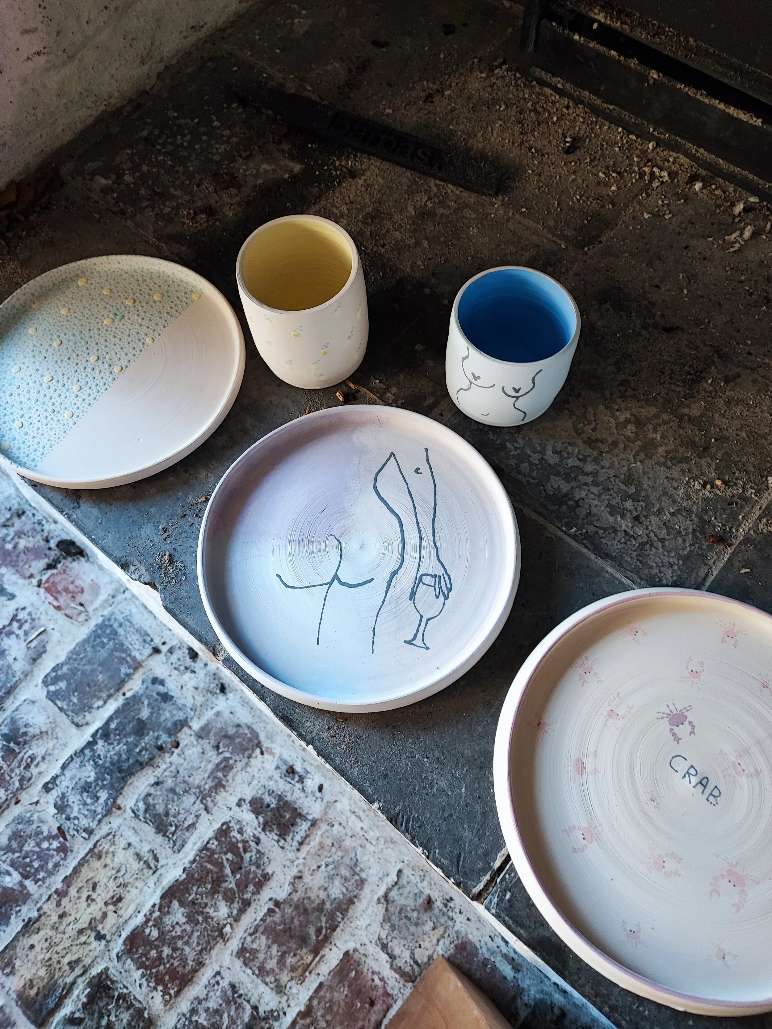 Keramische schalen met decoraties en bekers, een met een gele binnenkant en een met een blauwe. Op de schalen staan tekeningen van een figuur met een wijnglas en kleine krabben en het woord 'CRAB'. Ze staan op een stenen vloer.