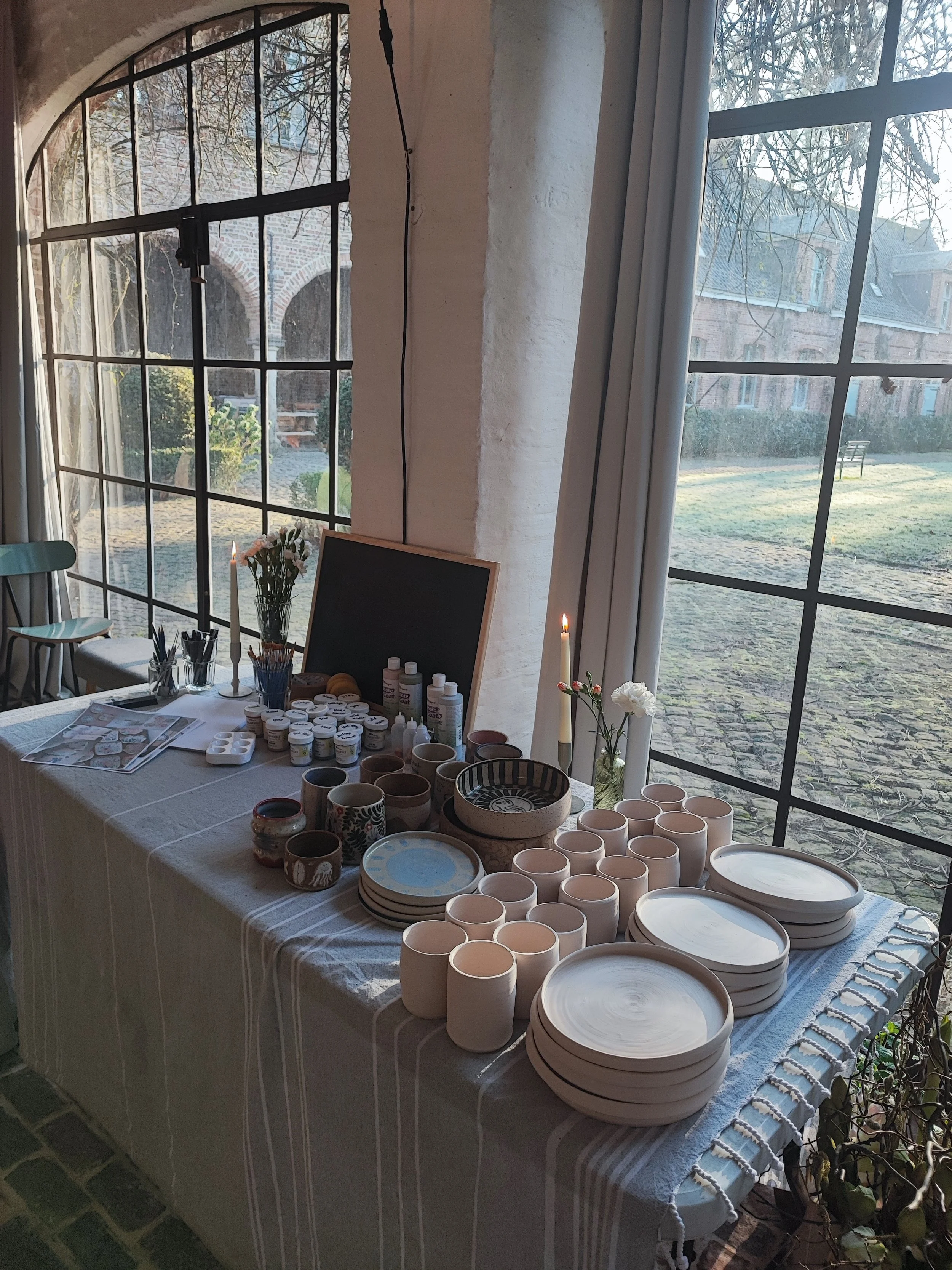 Tafel met keramische items, verfmaterialen en bloemen bij een raam, artistieke omgeving.