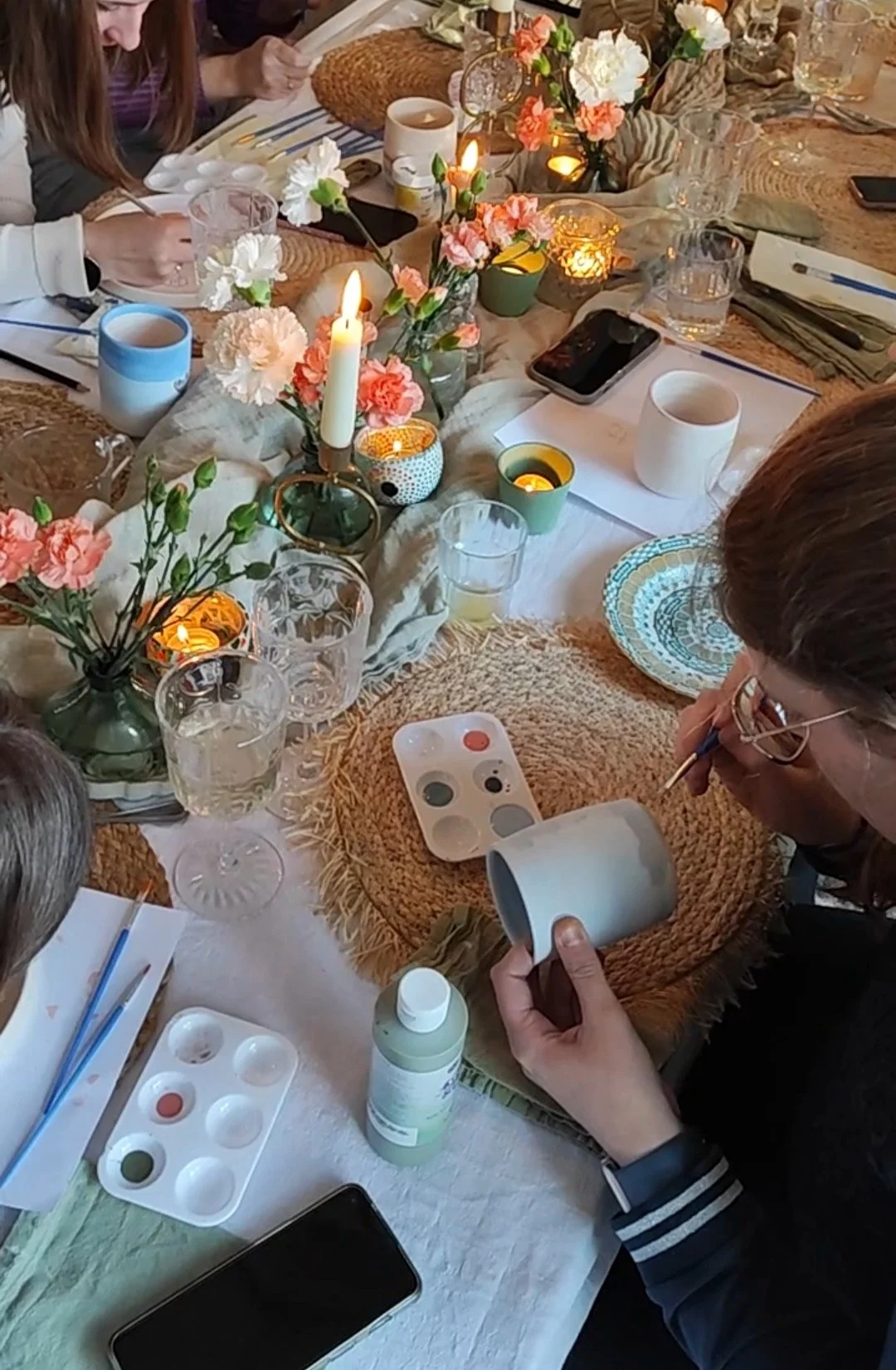 Een groep mensen schildert keramiek met verfpaletten en penselen aan een tafel vol decoraties zoals bloemen, kaarsen en glazen.