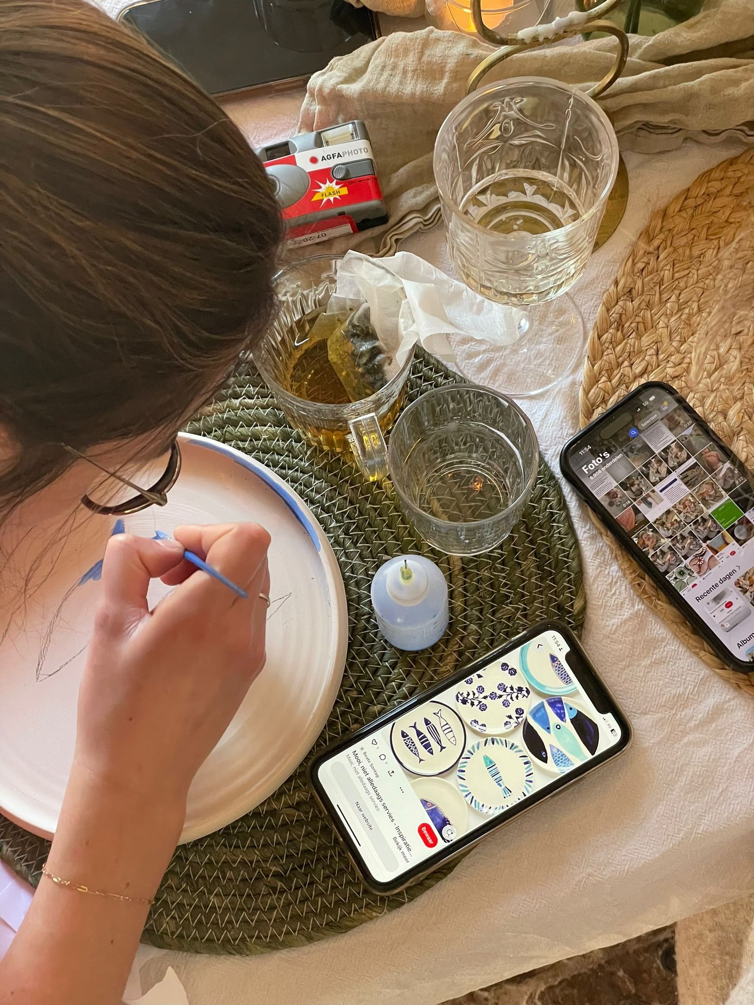 Persoon schildert een bord met een penseel, omringd door kopjes, een telefoon, en een rolletje film op tafel.