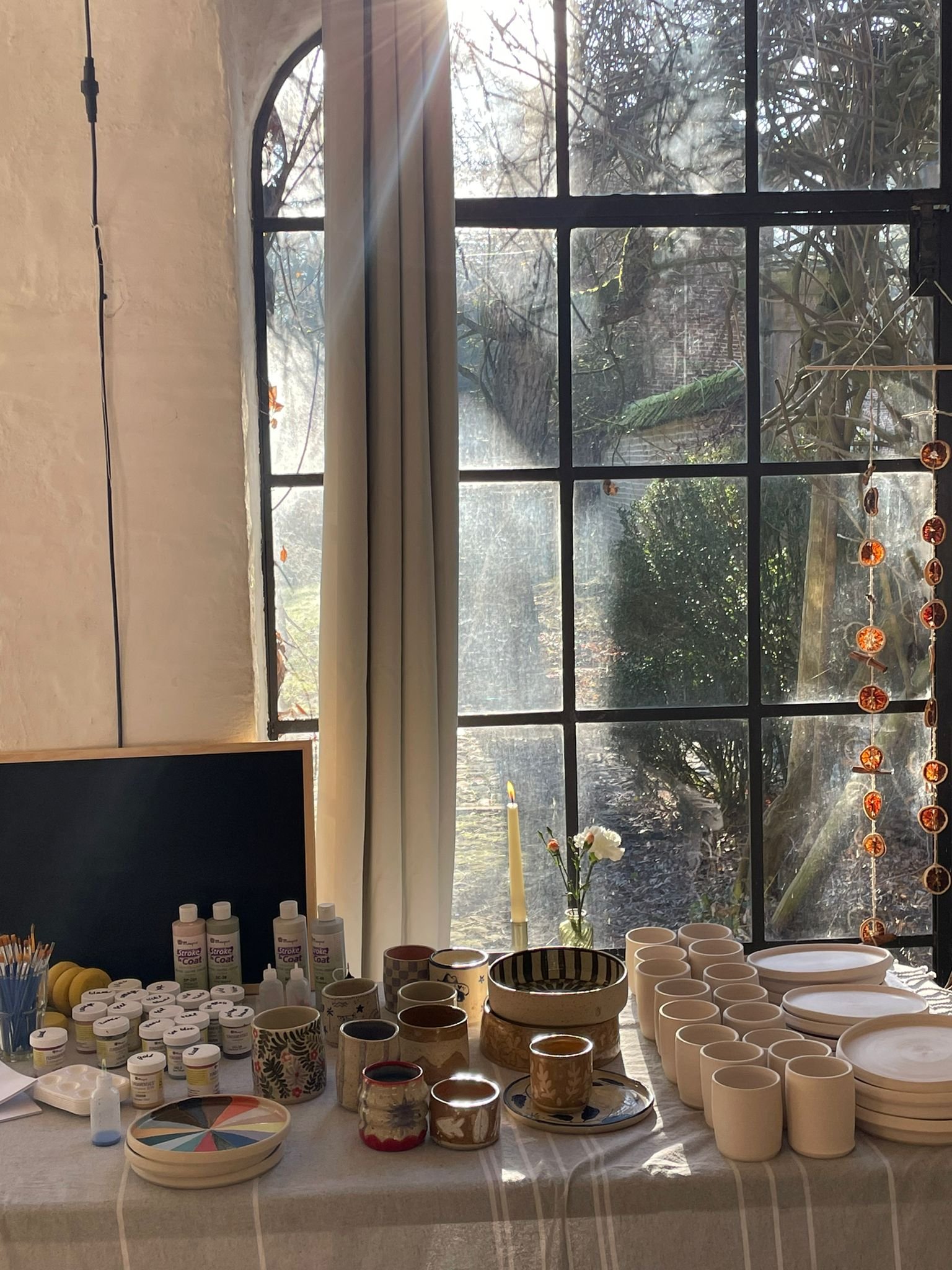Tafel met keramiekbenodigdheden en -producten bij een raam, zonlicht stroomt naar binnen; diverse potten, schalen en verfmaterialen, met gedroogde sinaasappelschijfjes en een enkele bloem in een vaas.