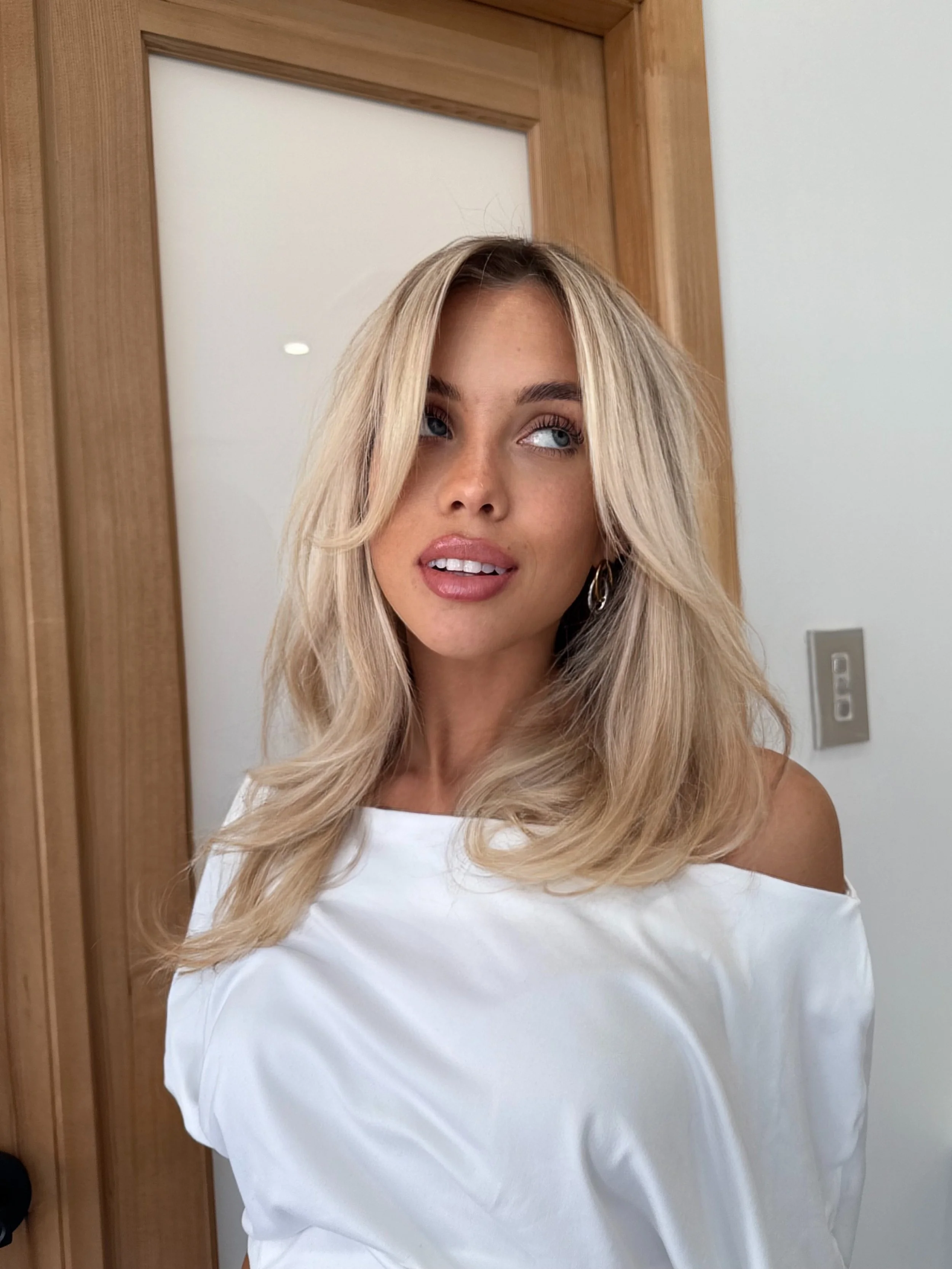 A young woman with blonde, wavy hair and blue eyes taking a selfie indoors. She is wearing a white top with shoulder cutouts and gold earrings, standing in front of a wooden door and a white wall.