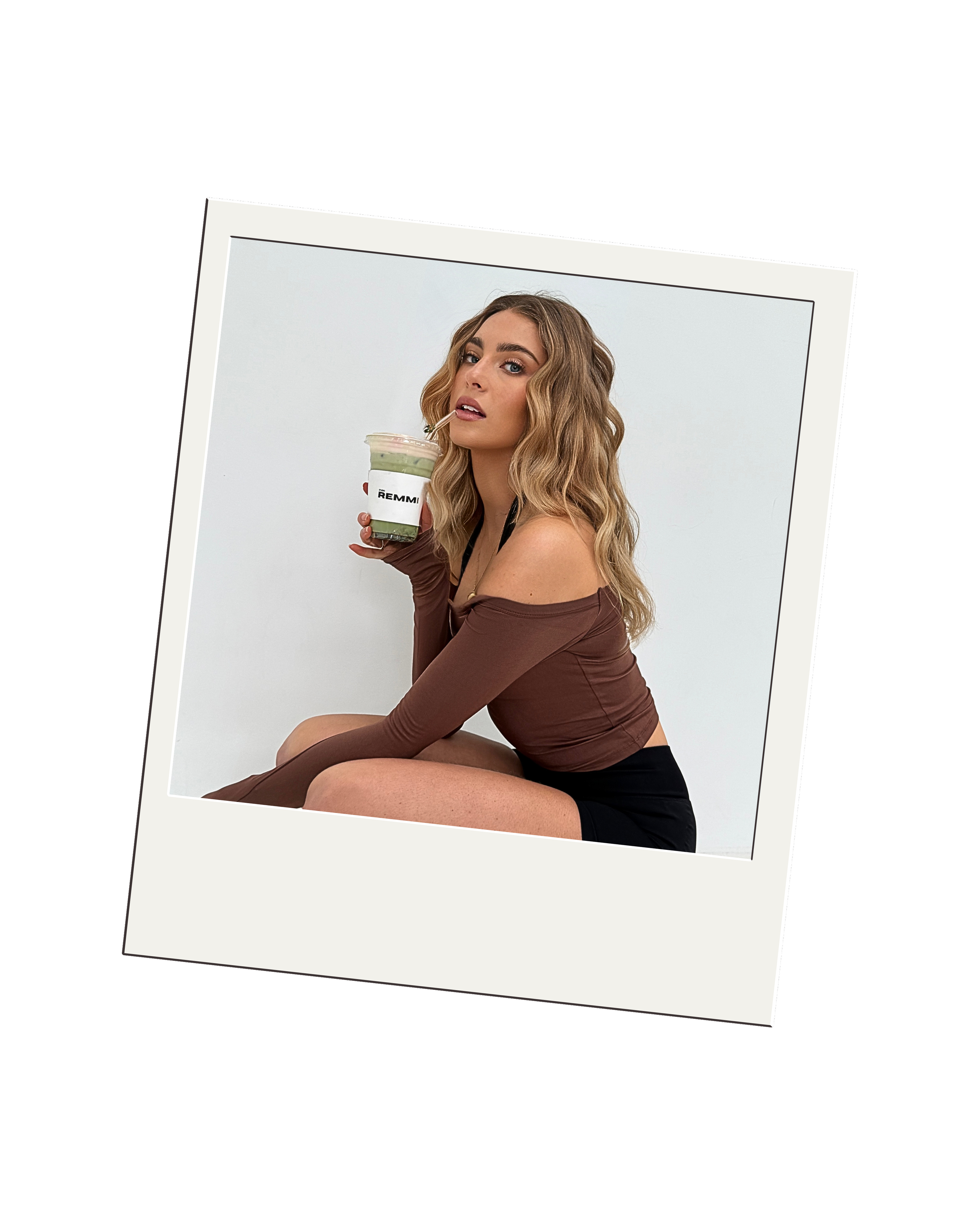 A young woman wearing a brown off-shoulder top and black shorts sitting against a white background, holding a cup of iced coffee or tea with a straw, and looking at the camera.
