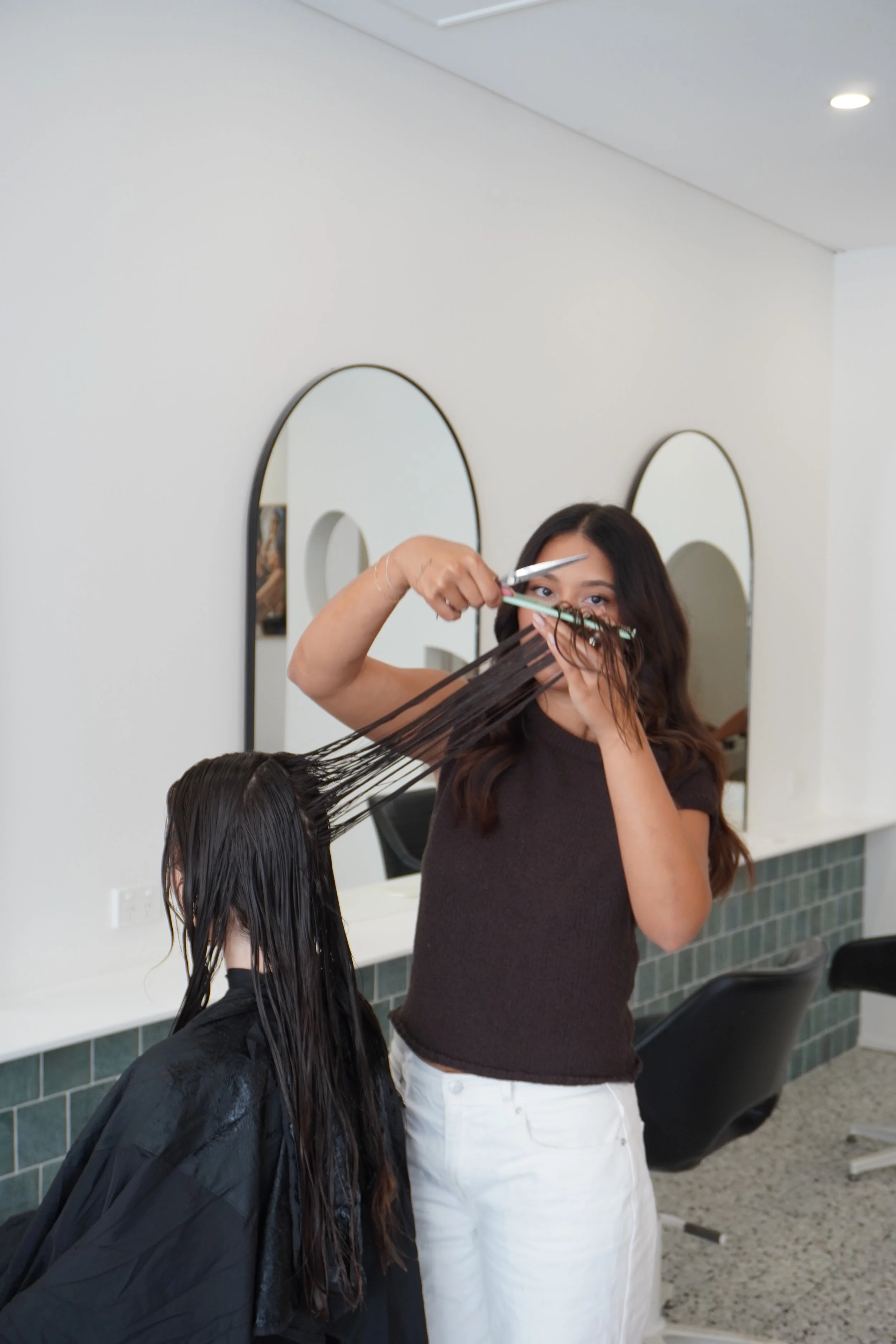 A hairdresser is cutting a woman's long, dark hair at a salon, with mirrors and chairs visible in the background.