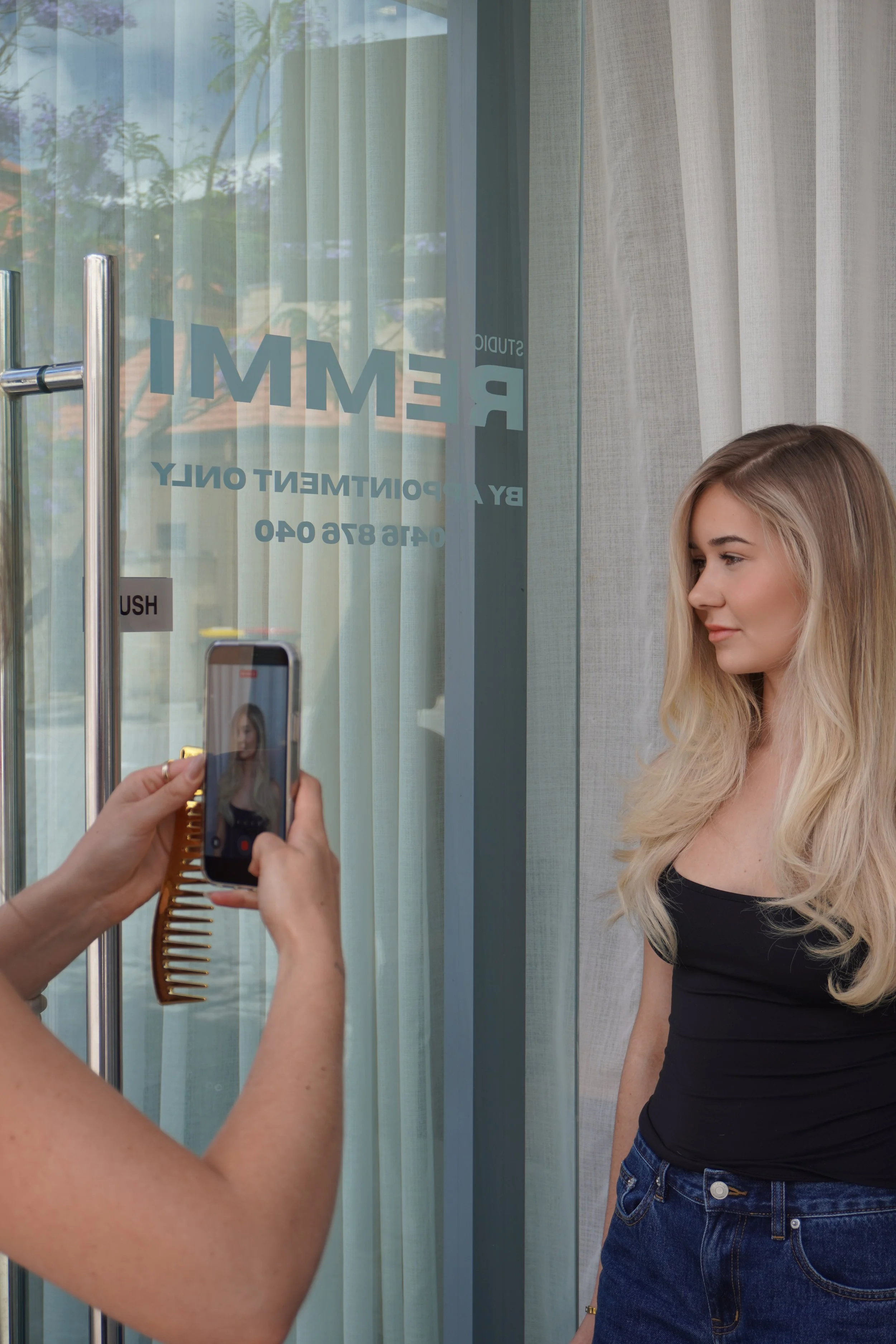 A woman with long blonde hair taking a photo of a young woman with long blonde hair standing near a glass door with the word 'REMI' and business contact info on it.
