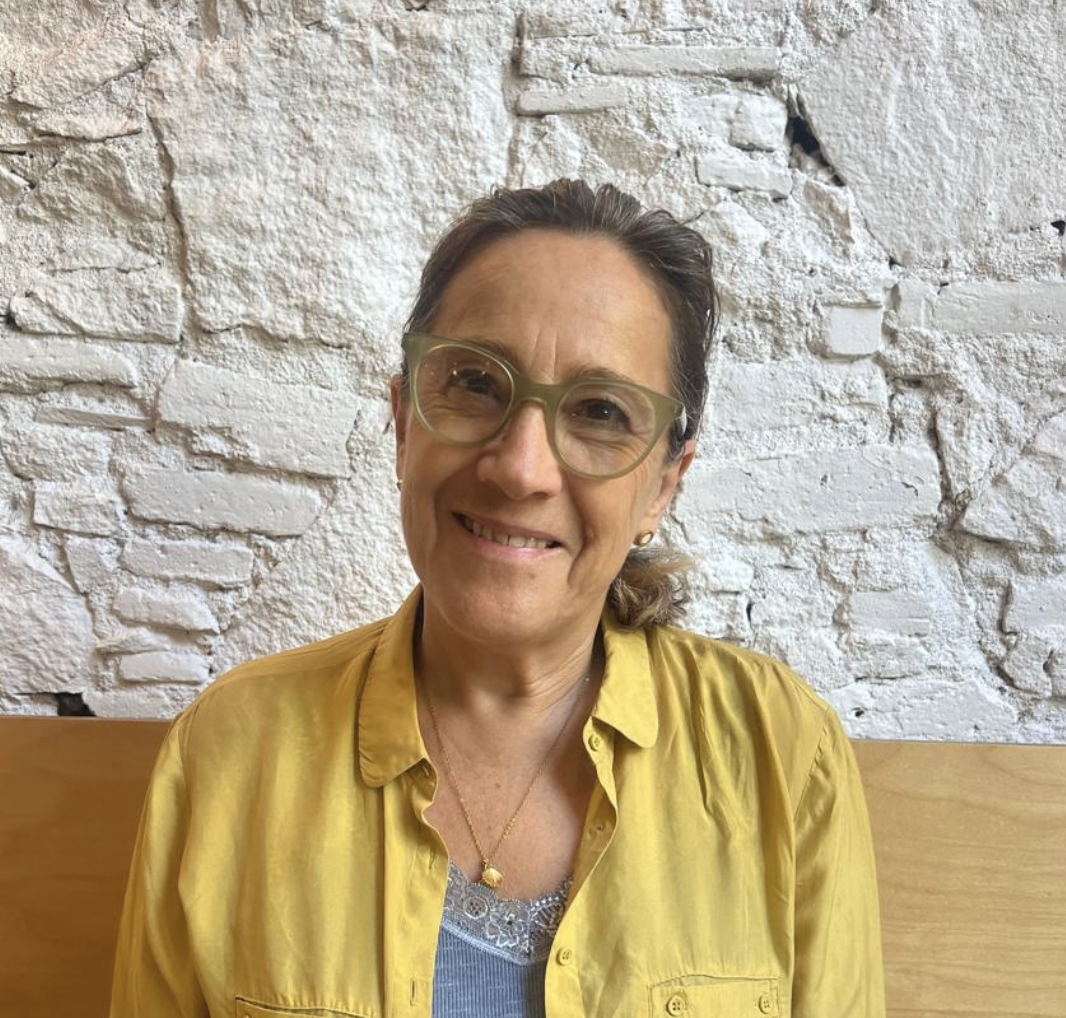 Mujer sonriente con gafas de pasta y blusa amarilla, sentada frente a una pared de ladrillos blancos.