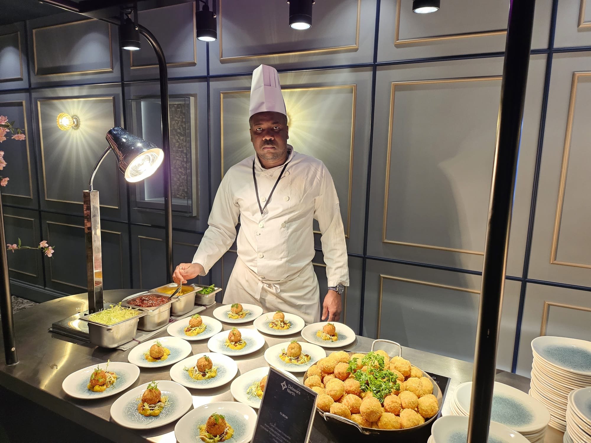 A chef in a white uniform and hat stands at a buffet table with various plated appetizers. The background features a decorative gray wall with gold trim, and there is warm lighting above.