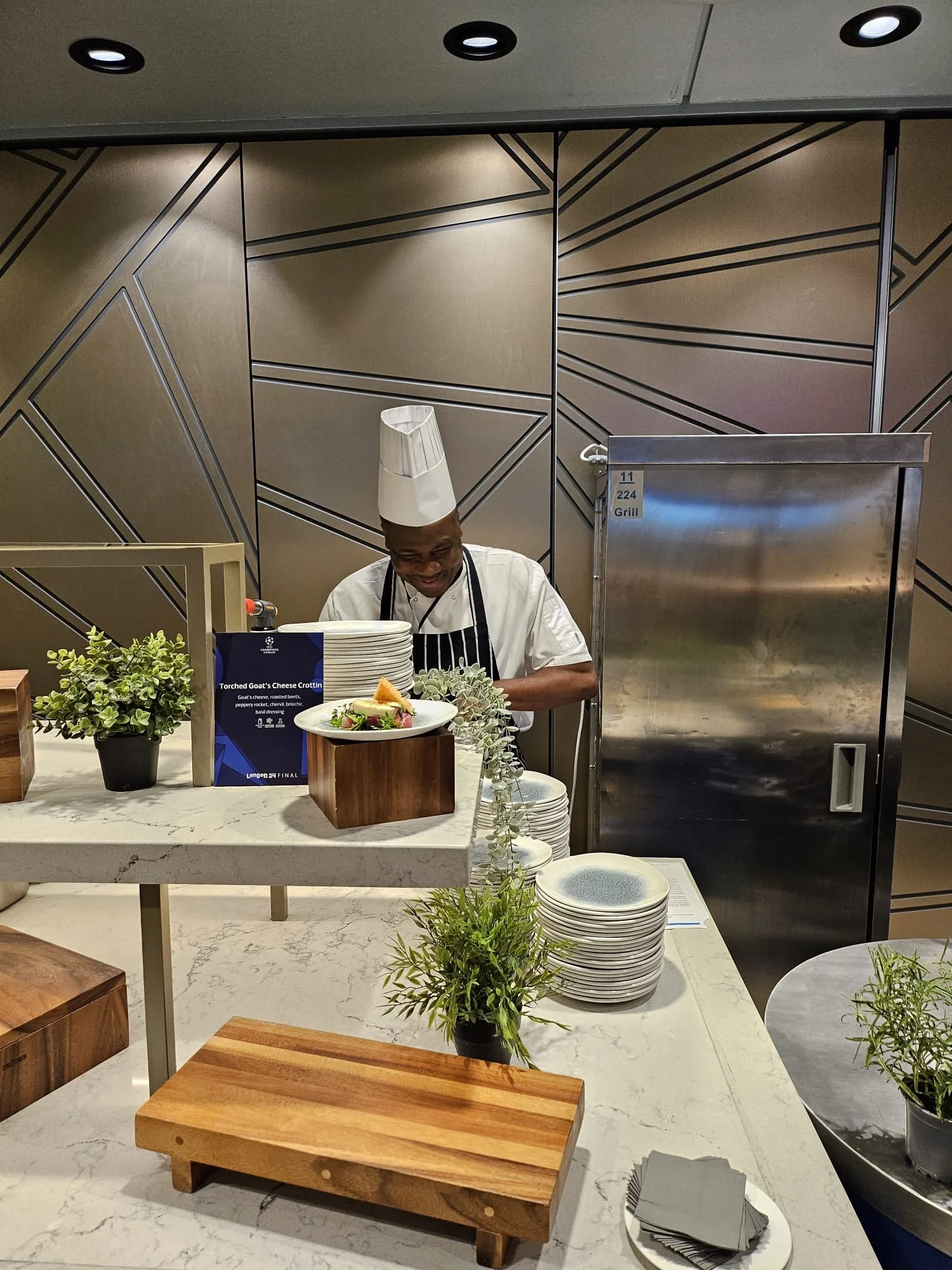 Chef smiling behind a counter with plates, plants, and a sign, in a modern restaurant setting.