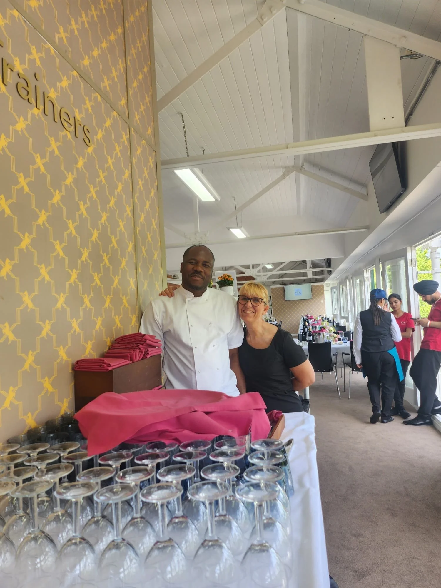 Two people, a man in a chef's coat and a woman in a black shirt, standing behind a table with upside-down wine glasses, inside a restaurant or event space.