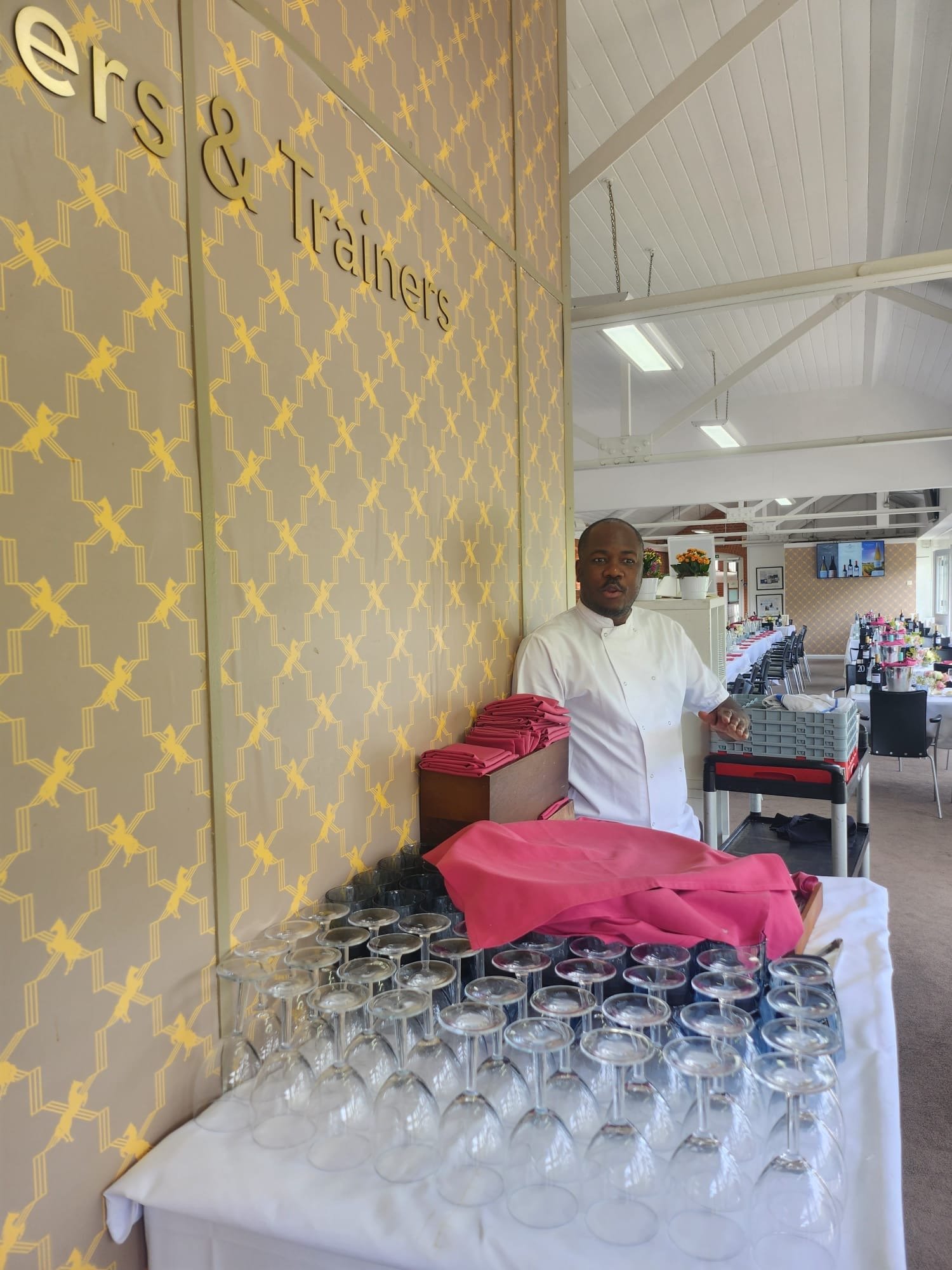 A chef standing next to a table set for an event in a decorated dining area, with glasses laid upside down and neatly folded napkins.