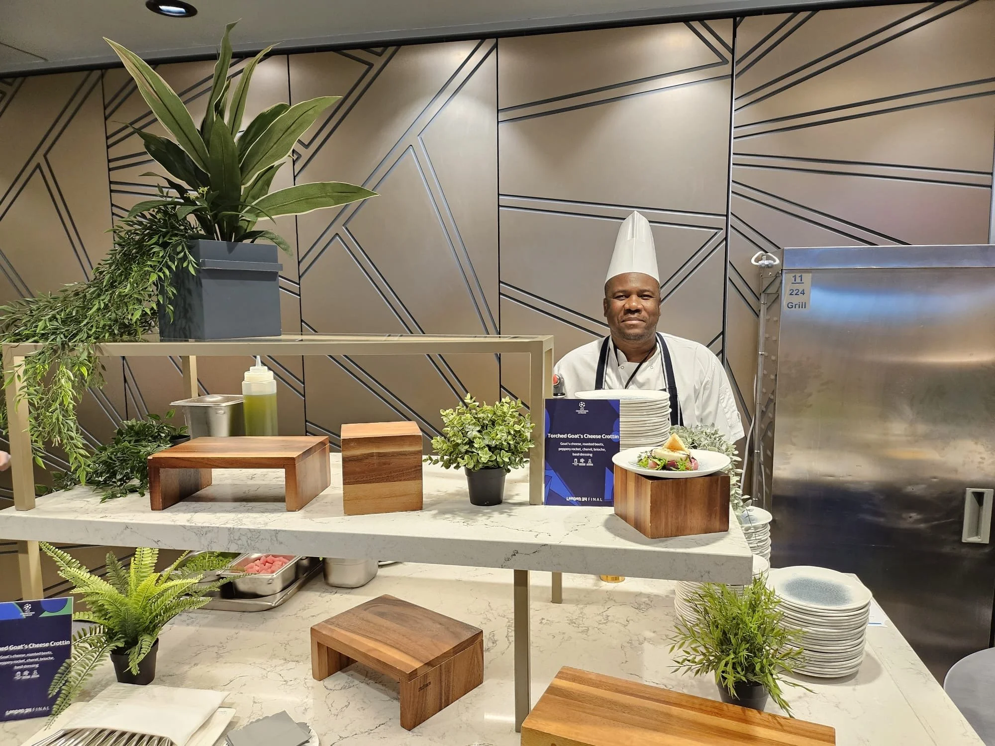 Chef standing behind a kitchen counter decorated with potted plants, wooden blocks, plates, and a sign promoting goat cheese crottin at a culinary event.