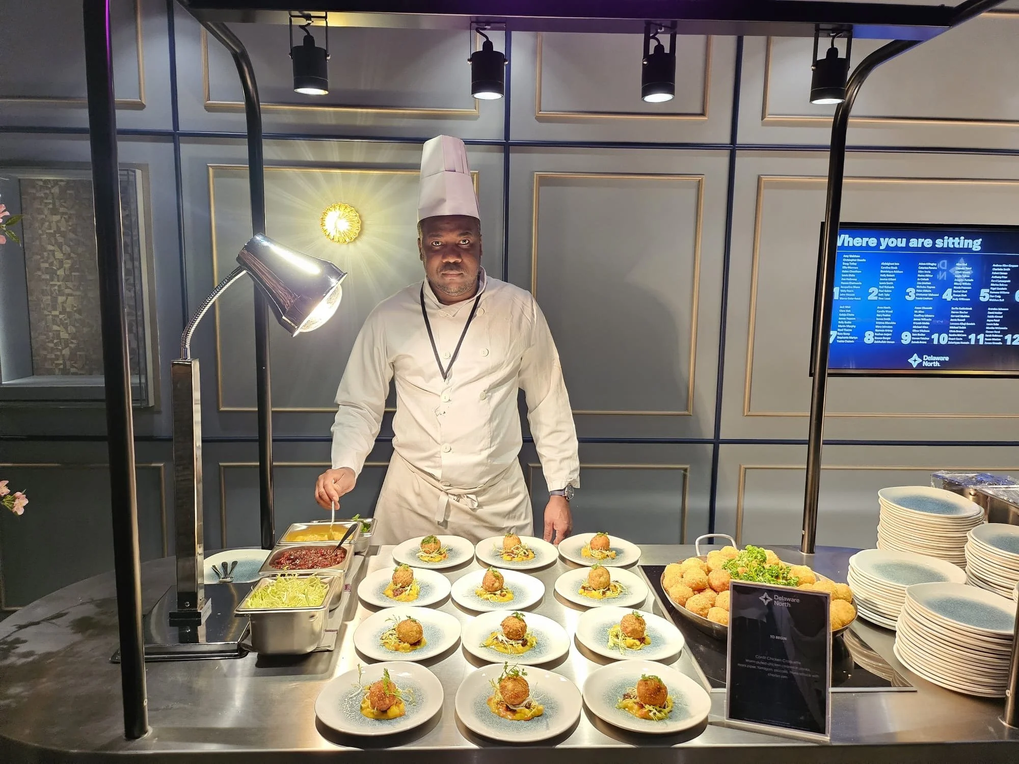 A chef wearing a white uniform and hat stands behind a buffet table with plates of food, salad, and breaded appetizers in a restaurant or hotel setting.