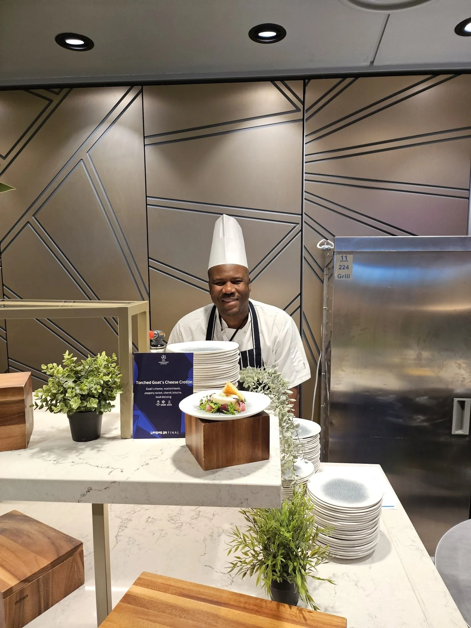 Chef standing behind a counter, smiling, in a modern kitchen setting with plates, plants, and a sign showcasing a dish called 'Torched Goat's Cheese Crottin'.