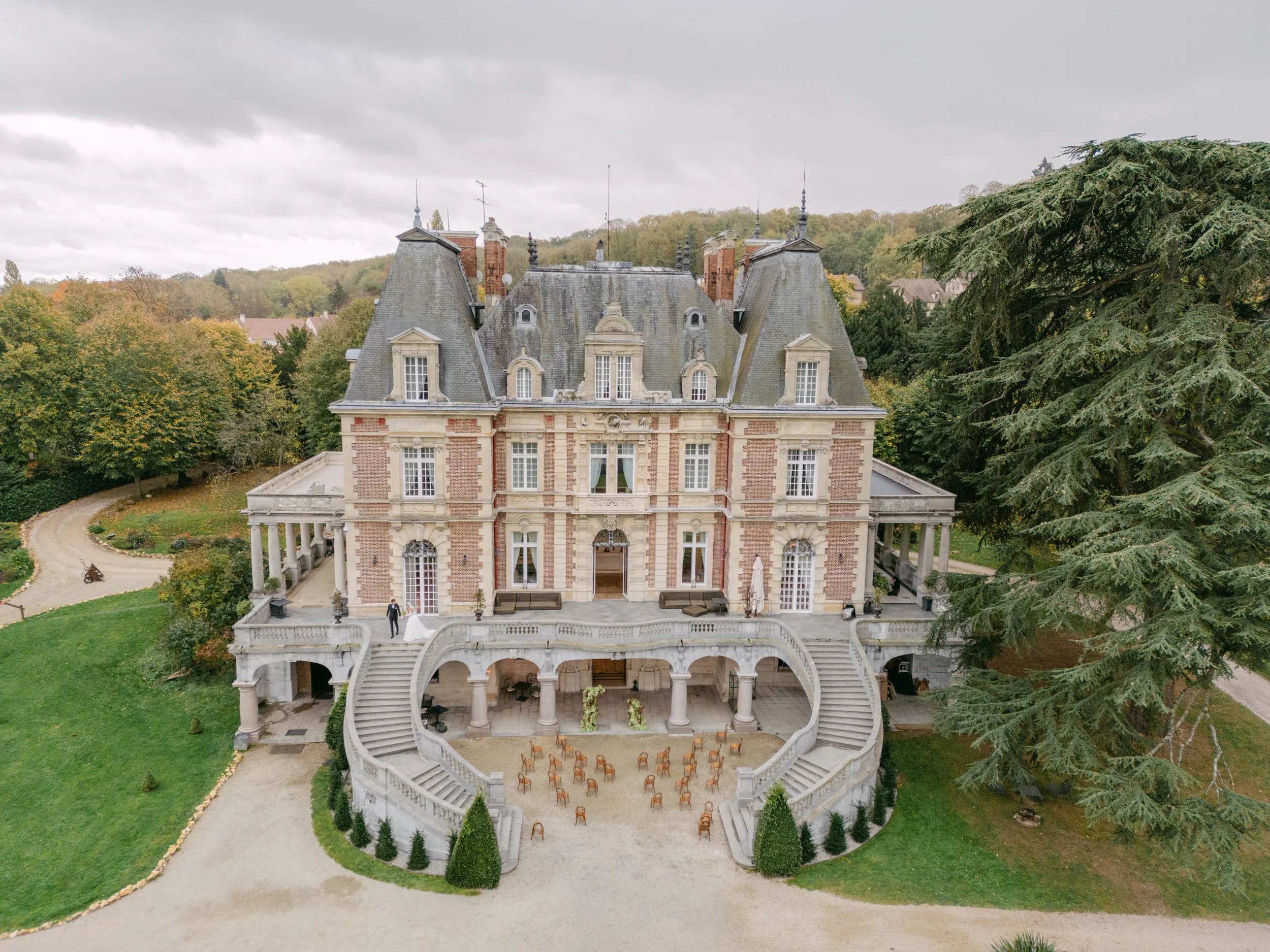 Ein großes Herrenhaus im französischen Stil mit steilen Dächern, mehreren Schornsteinen und einer schönen Fassade, umgeben von einem gepflegten Garten, große Bäume und eine Zufahrtsstraße.