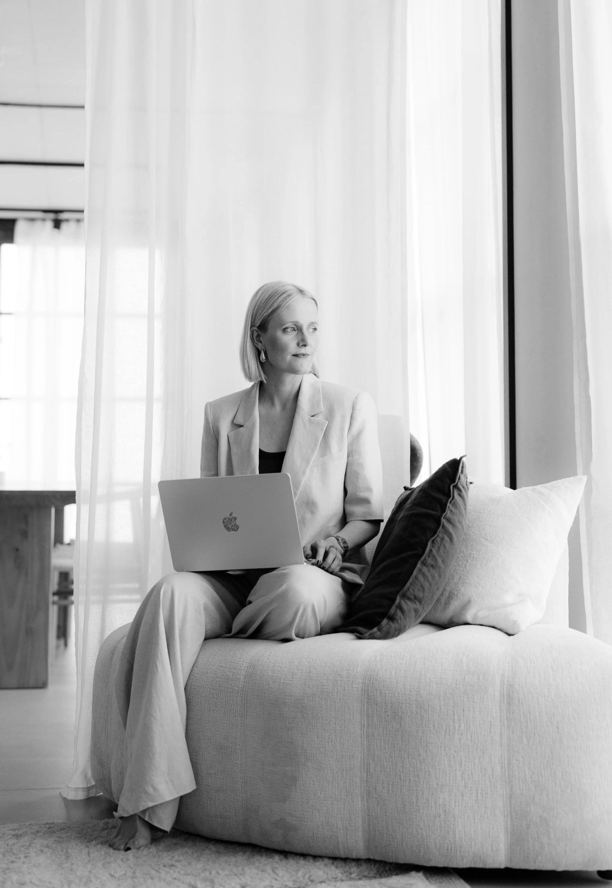 Frau sitzt auf einem Sofa, schaut aus dem Fenster, trägt eine beige Jacke und weiße Hosen, arbeitet an einem Laptop.