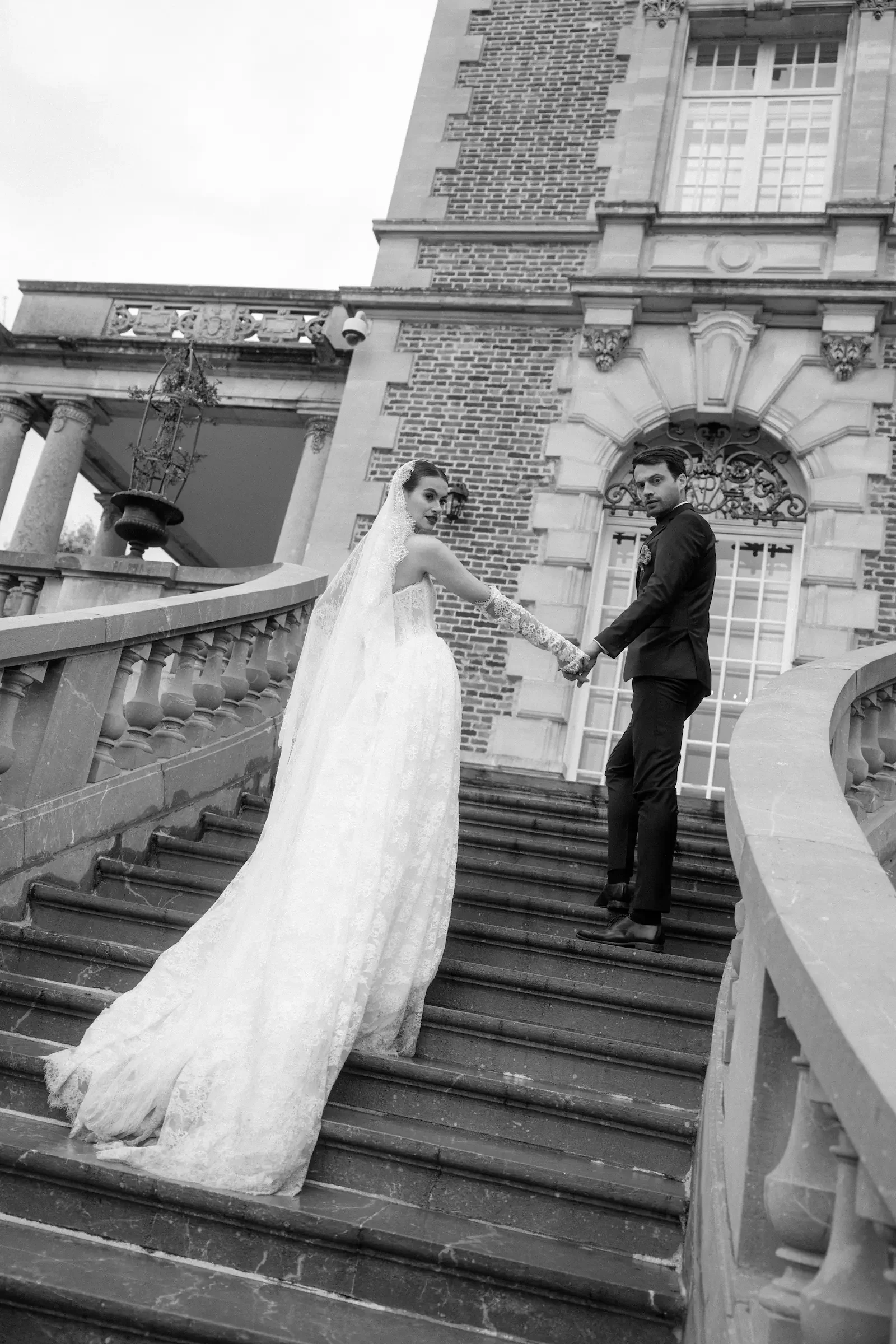 Ein Brautpaar steigt die Treppe zu einem historischen Gebäude mit Stein und Ziegelsteinfassade, die Frau trägt ein langes Brautkleid mit Schleier, der Mann ein dunklen Anzug.