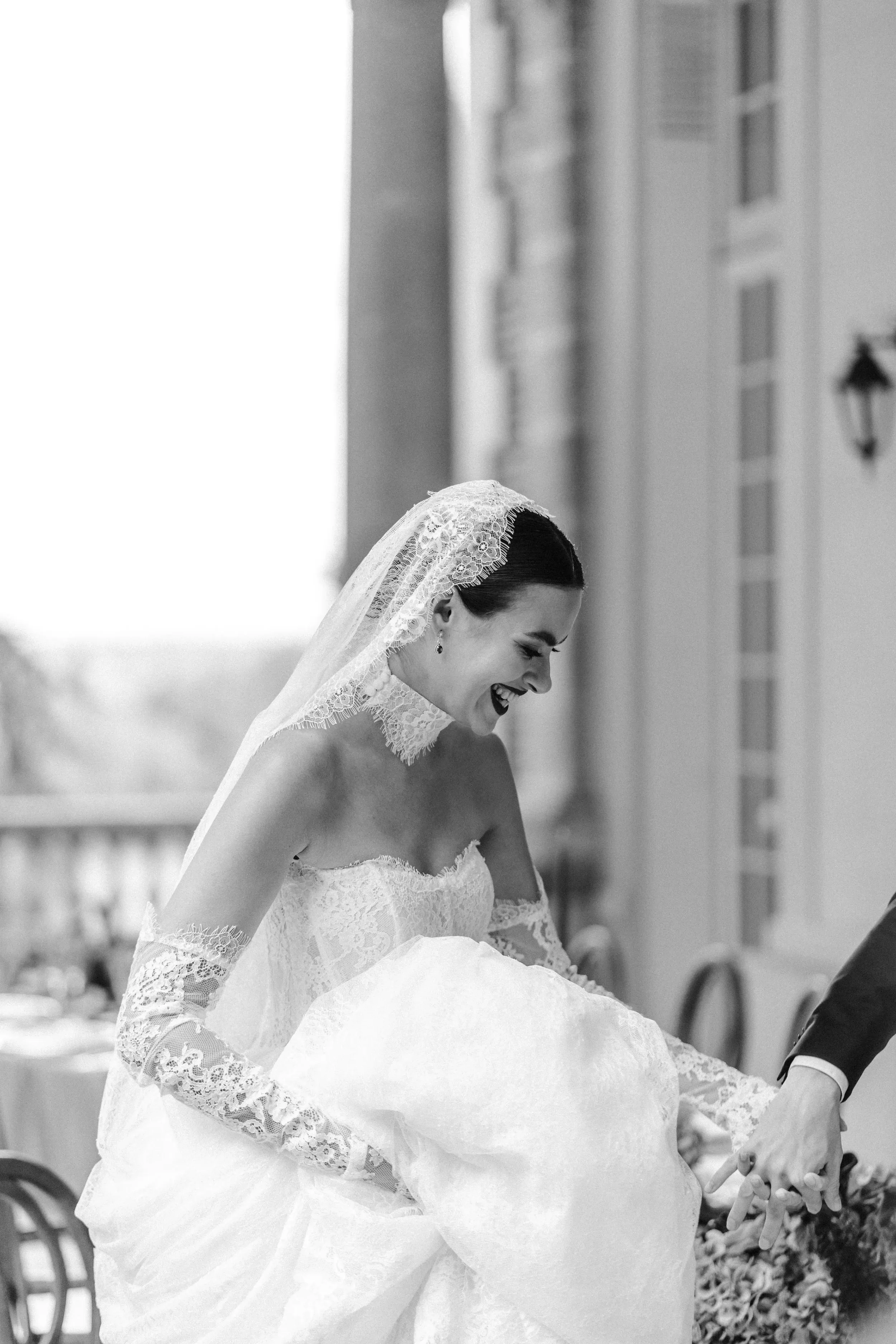 Braut in Hochzeitskleid und Schleier lacht, während sie gehalten wird, bei einer Hochzeit im Freien.