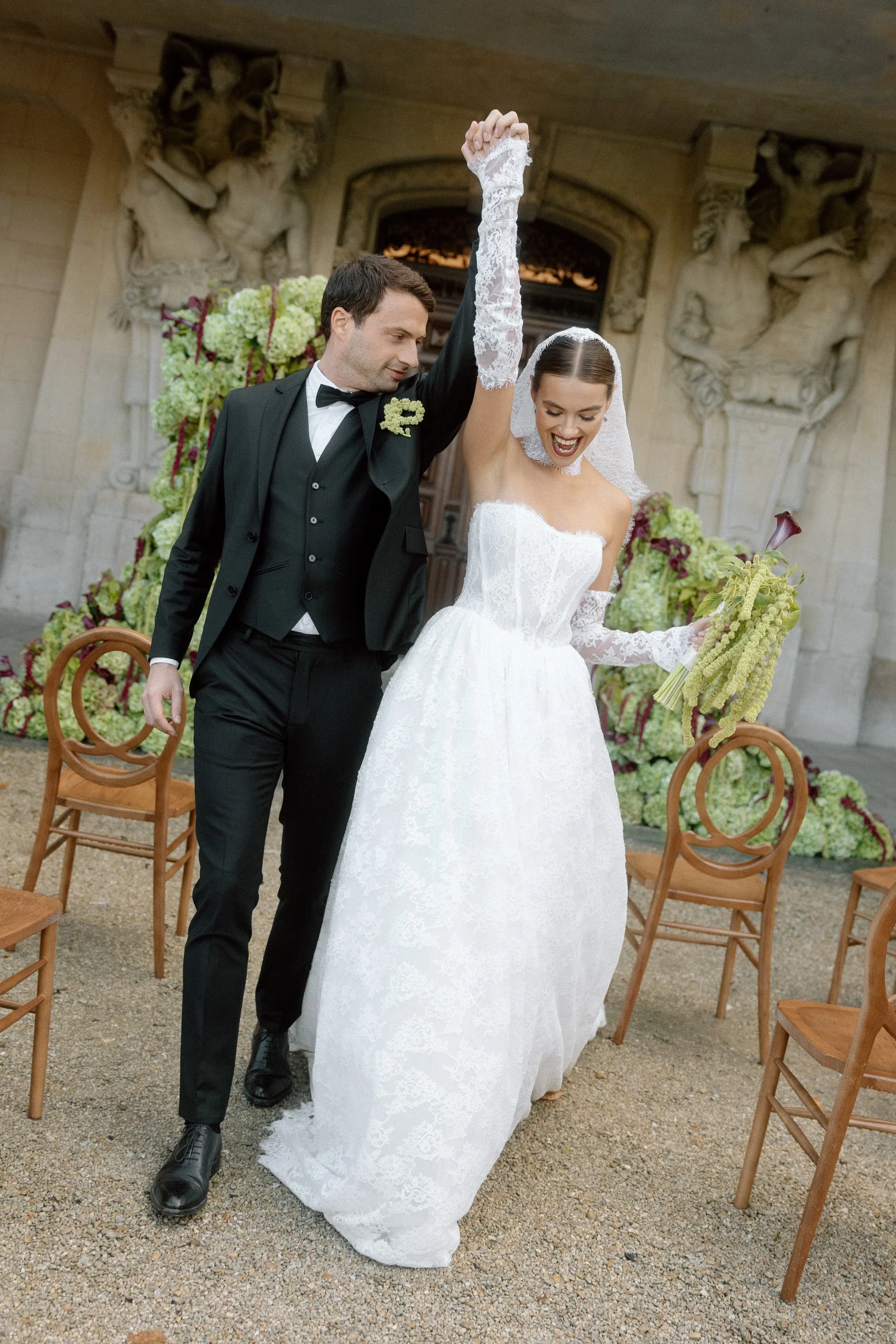 Ein frisch verheiratetes Paar tanzt und lacht bei einer Hochzeit im Freien, umgeben von Stühlen und Dekorationen mit Blumen, vor einer historischen Gebäude mit Skulpturen.