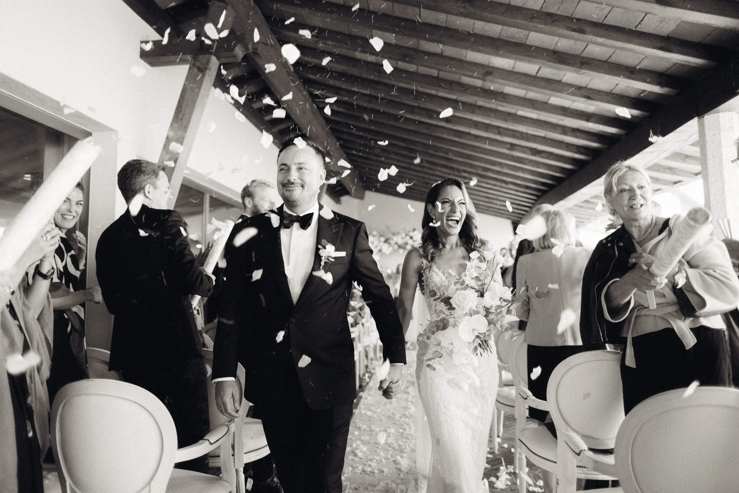 Ein Hochzeitsfoto in Schwarz-Weiß mit einem lachenden Brautpaar, das gemeinsam durch einen Konfettiregen läuft, umgeben von fröhlichen Gästen.