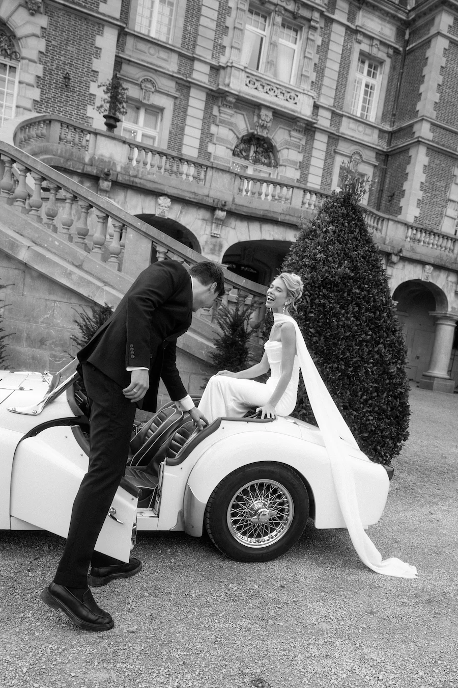 Ein Brautpaar in formeller Kleidung auf einem Oldtimer-Cabrio, vor einem historischen Gebäude, vermutlich bei einer Hochzeit.