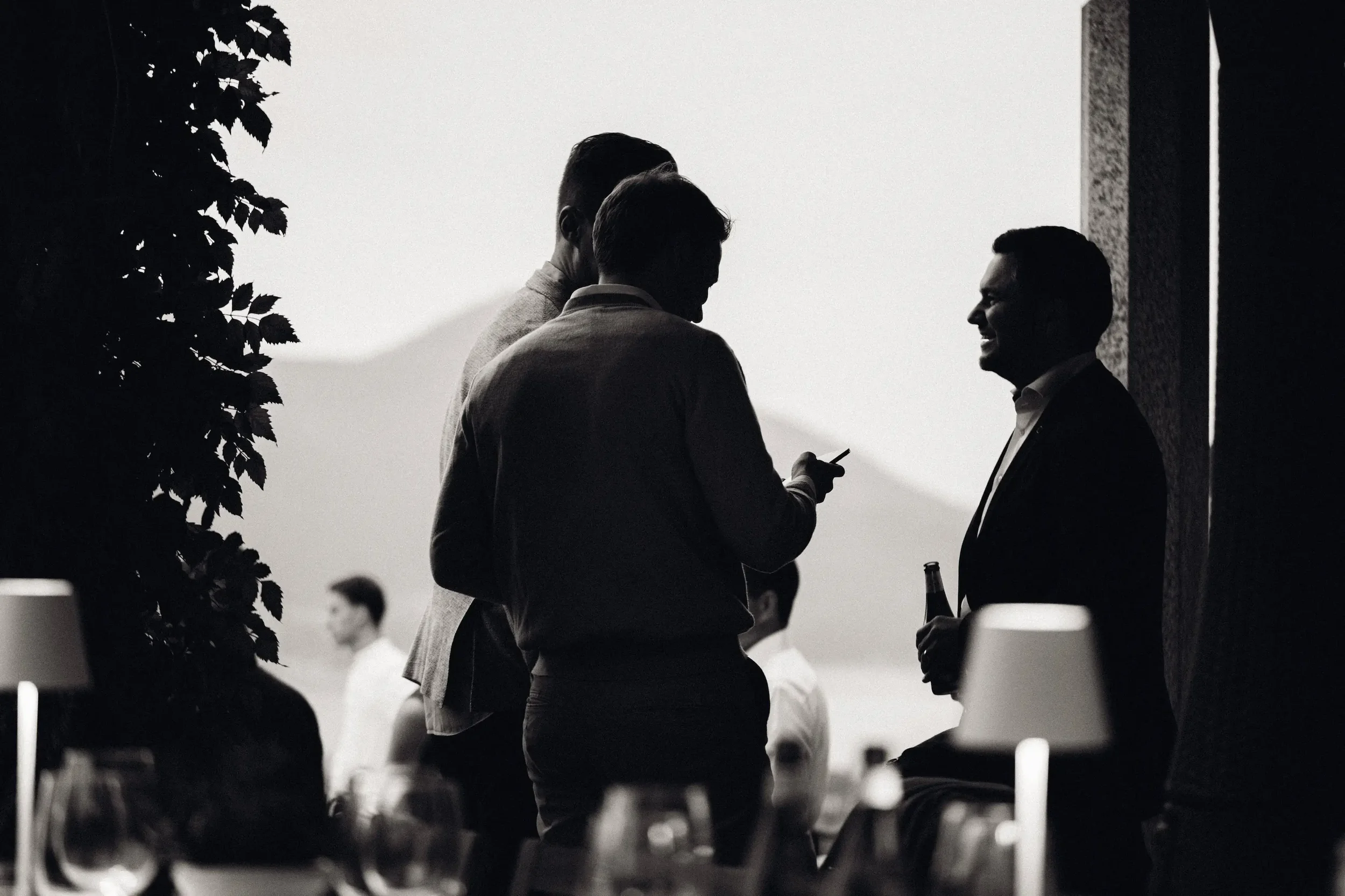 Schattenhafte Silhouetten von Männern in einem Gespräch bei einem Event, mit Tischen und Lampen im Vordergrund, im Hintergrund eine Berglandschaft