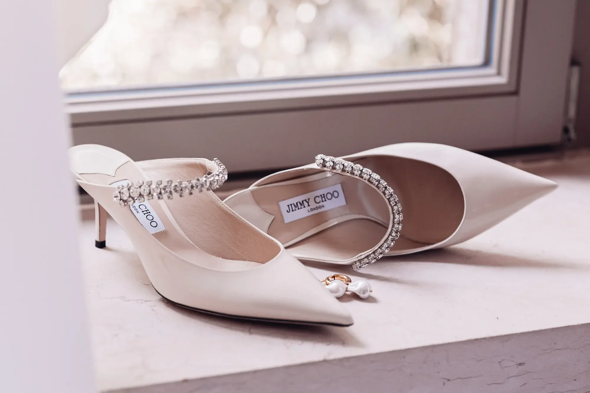 Weißes Paar High Heels von Jimmy Choo mit Schmuck am Knöchel, auf einem Fensterbank neben einem Fenster