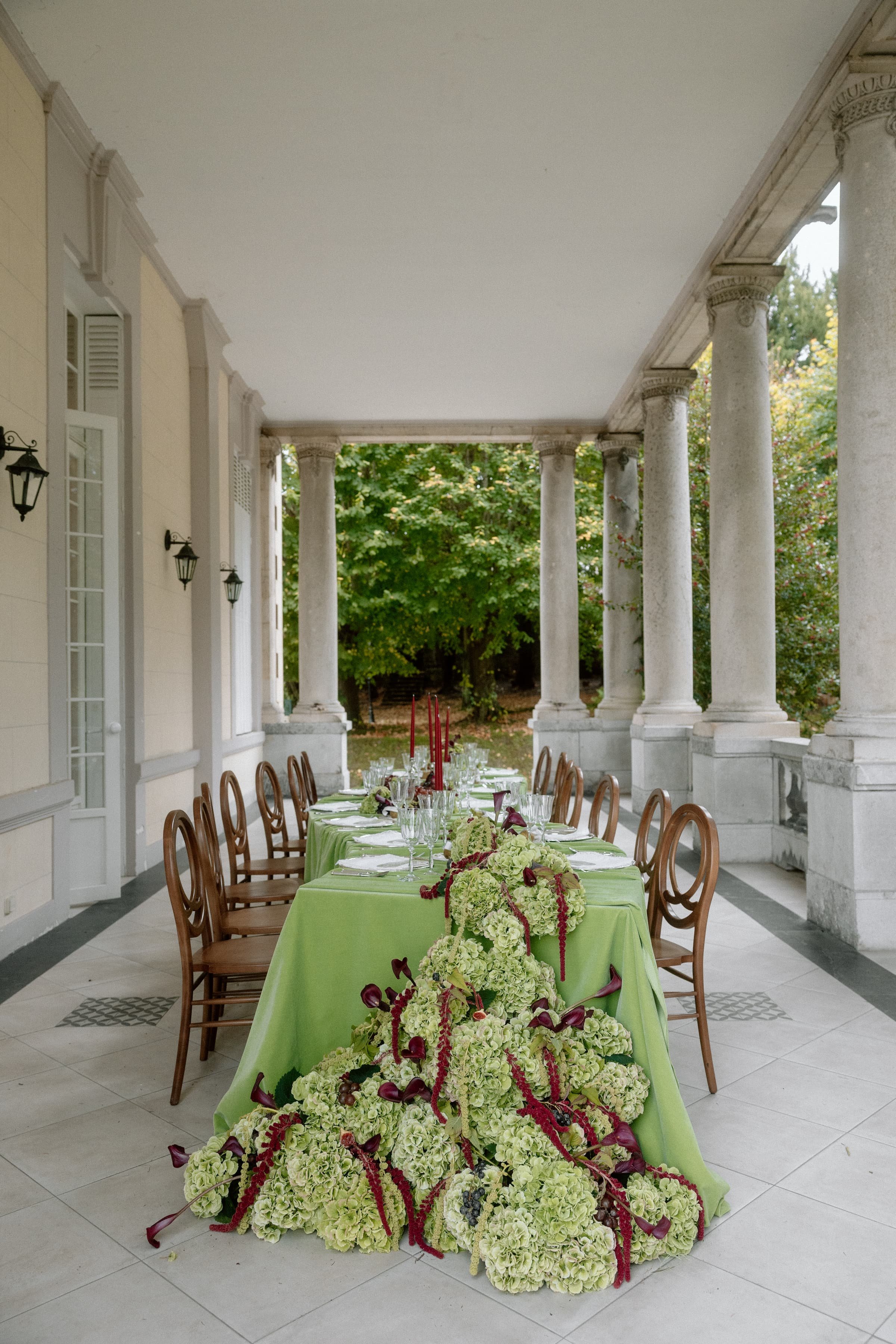 Ein festlich gedeckter Tisch mit grüner Tischdecke, festliche Blumenarrangements in Weiß und Violett, auf einer Veranda mit klassischen Säulen, im Hintergrund Bäume.