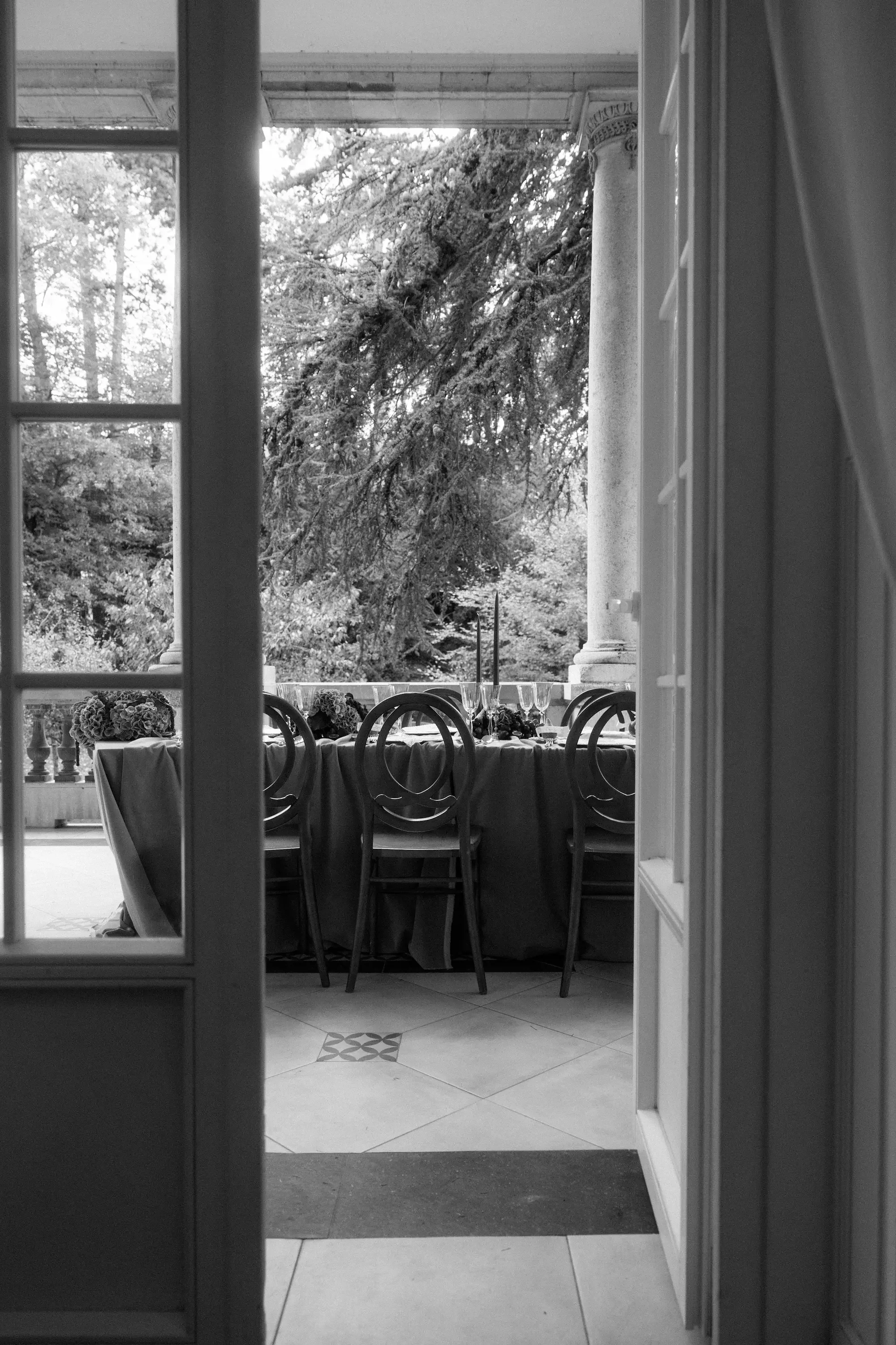 Ein eleganter Esstisch auf einer Terrasse mit Blick auf einen grünen Garten und eine große Baumkrone, gedeckt mit Geschirr, Gläsern und Blumen.
