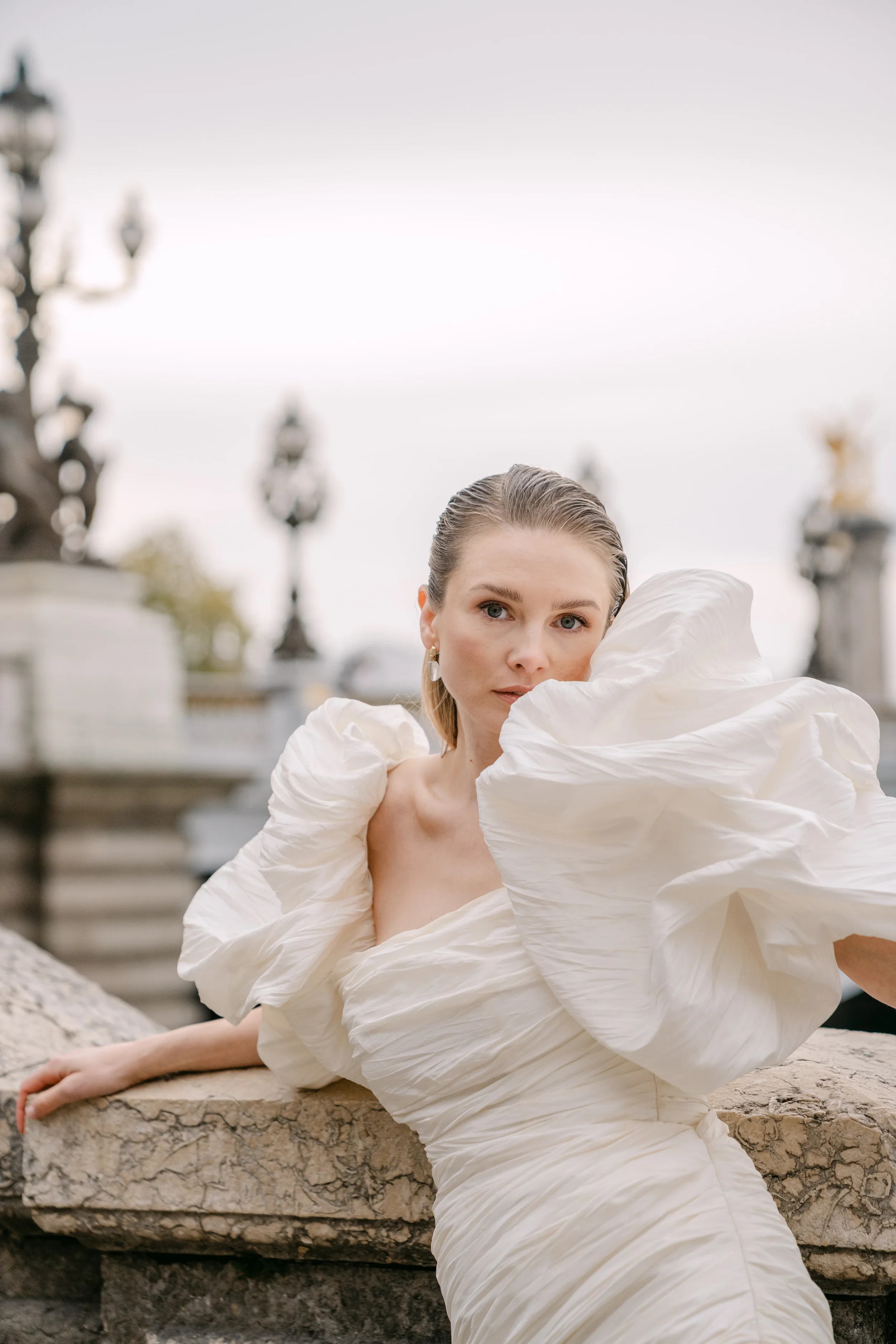 Eine Frau trägt ein elegantes, weißes, spitz zulaufendes Kleid mit großen, puffigen Ärmeln und posiert vor einer historischen, stimmungsvollen Kulisse mit Laternen und Statuen.