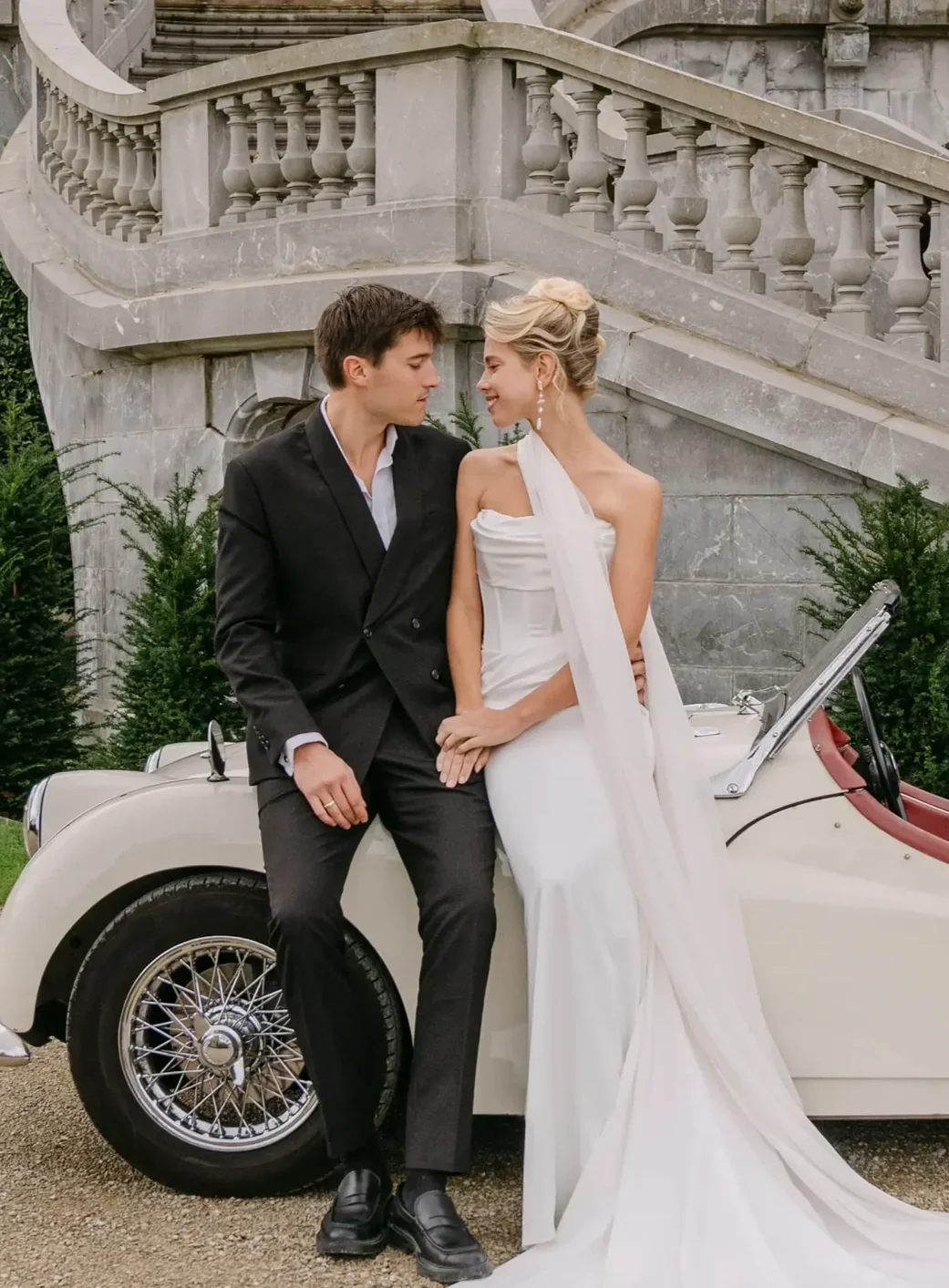 Ein junges Paar in formeller Kleidung, das vor einer historischen Steintreppe steht, mit einem weißen Oldtimer-Cabriolet, das im Vordergrund geparkt ist.