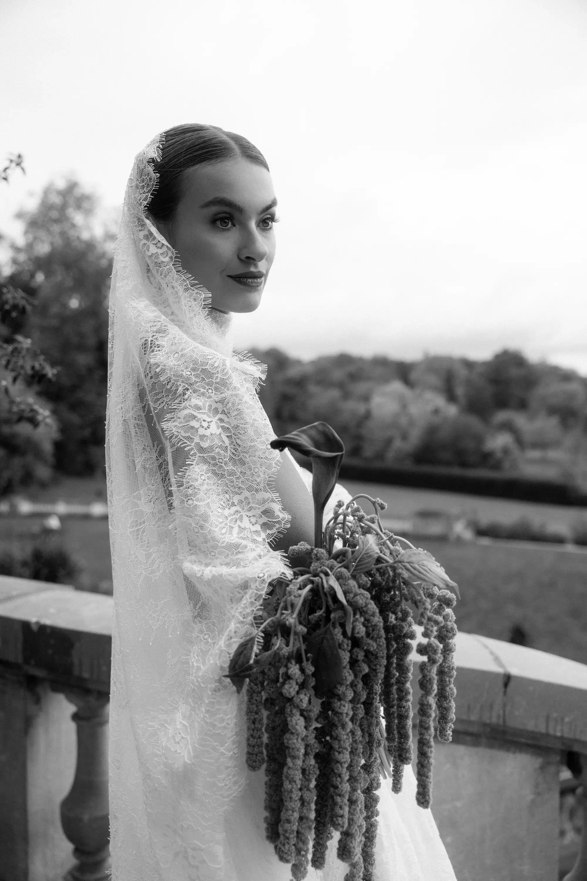 Schwarz-weiße Aufnahme einer jungen Frau in Brautkleid mit Schleier, die einen Blumenstrauß hält, im Freien mit Bäumen im Hintergrund.
