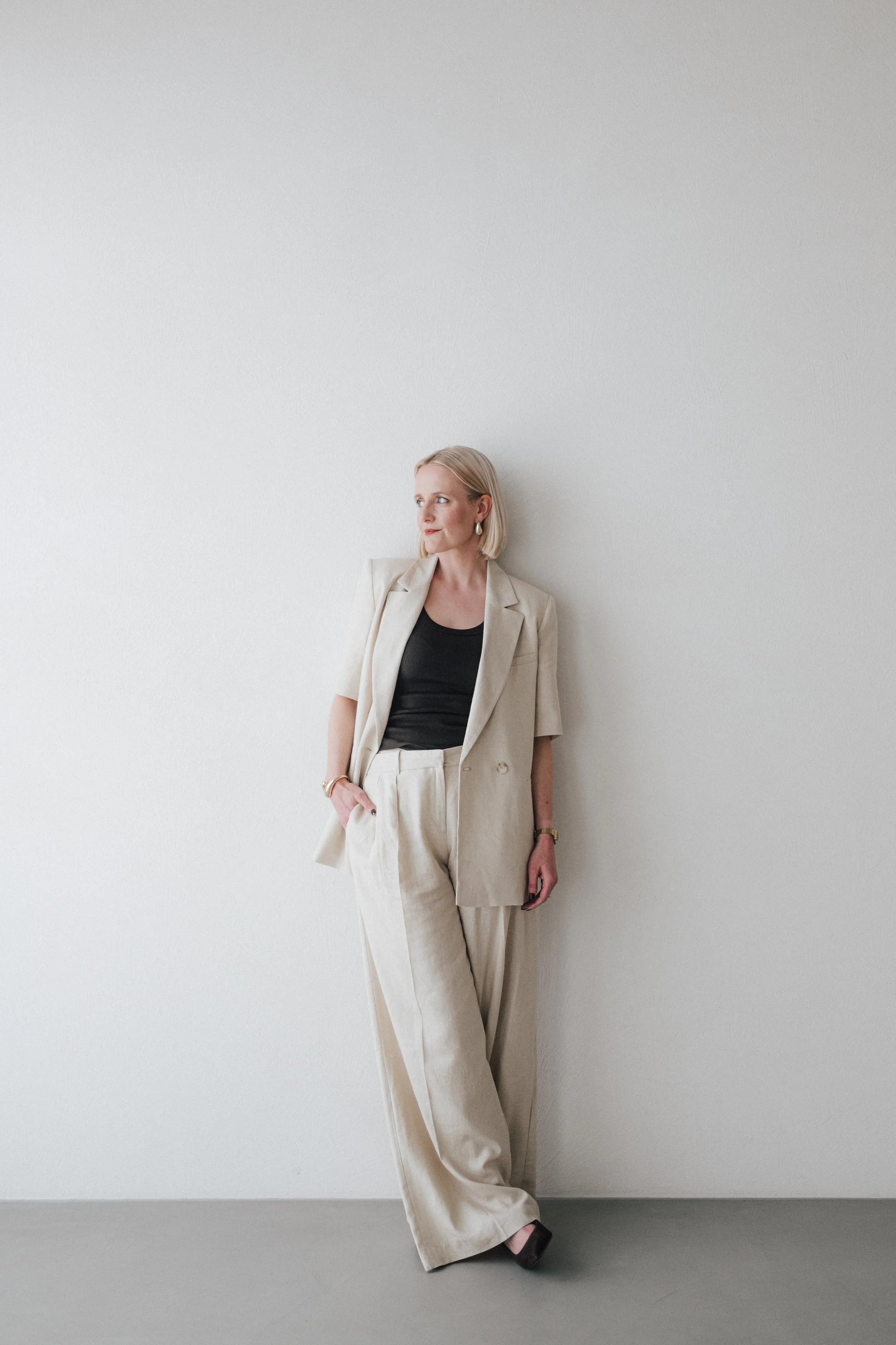 Frau in beige Anzug und schwarzem Top lehnt an einer weißen Wand, blickt nach links.