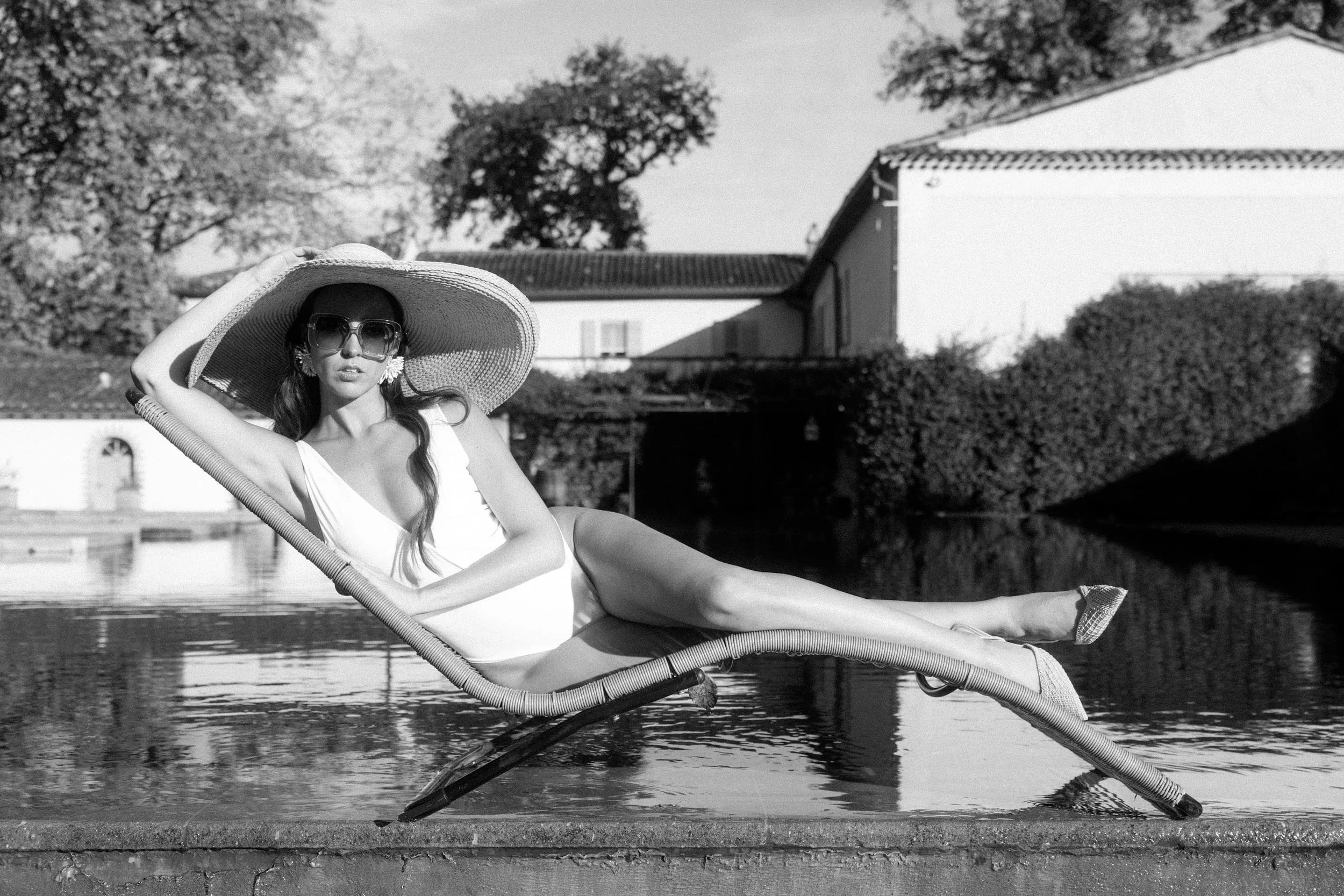 Frau in Sonnenbrille und großem Sonnenhut liegt auf einem Liegestuhl am Wasser, im Hintergrund Bäume und ein Gebäude, schwarz-weiß Aufnahme.