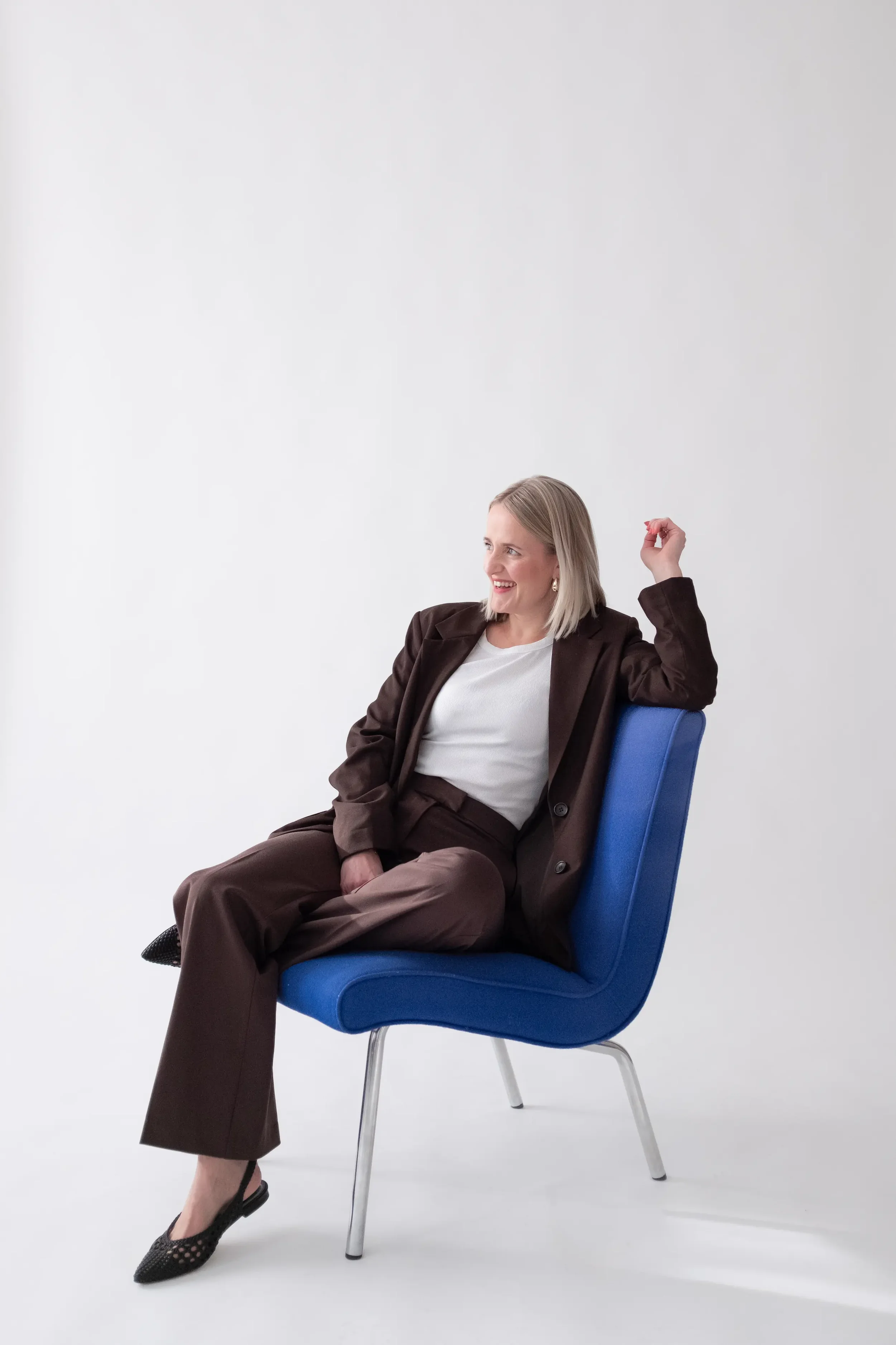 Frau in brauner Anzugsjacke und weißen Shirt sitzt lächelnd auf blauer Sessel vor weißen Hintergrund.