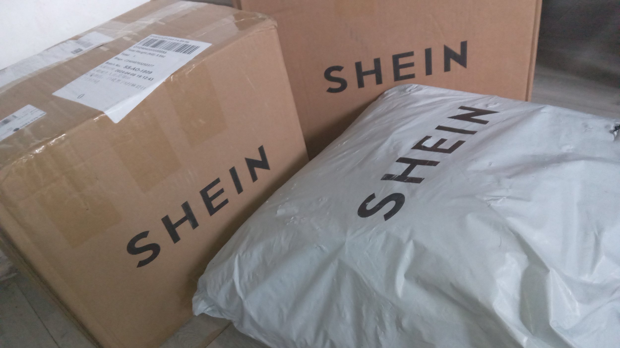 Three packages on a floor, one in white plastic wrapping and two in brown cardboard with black text 'SHEIN'.