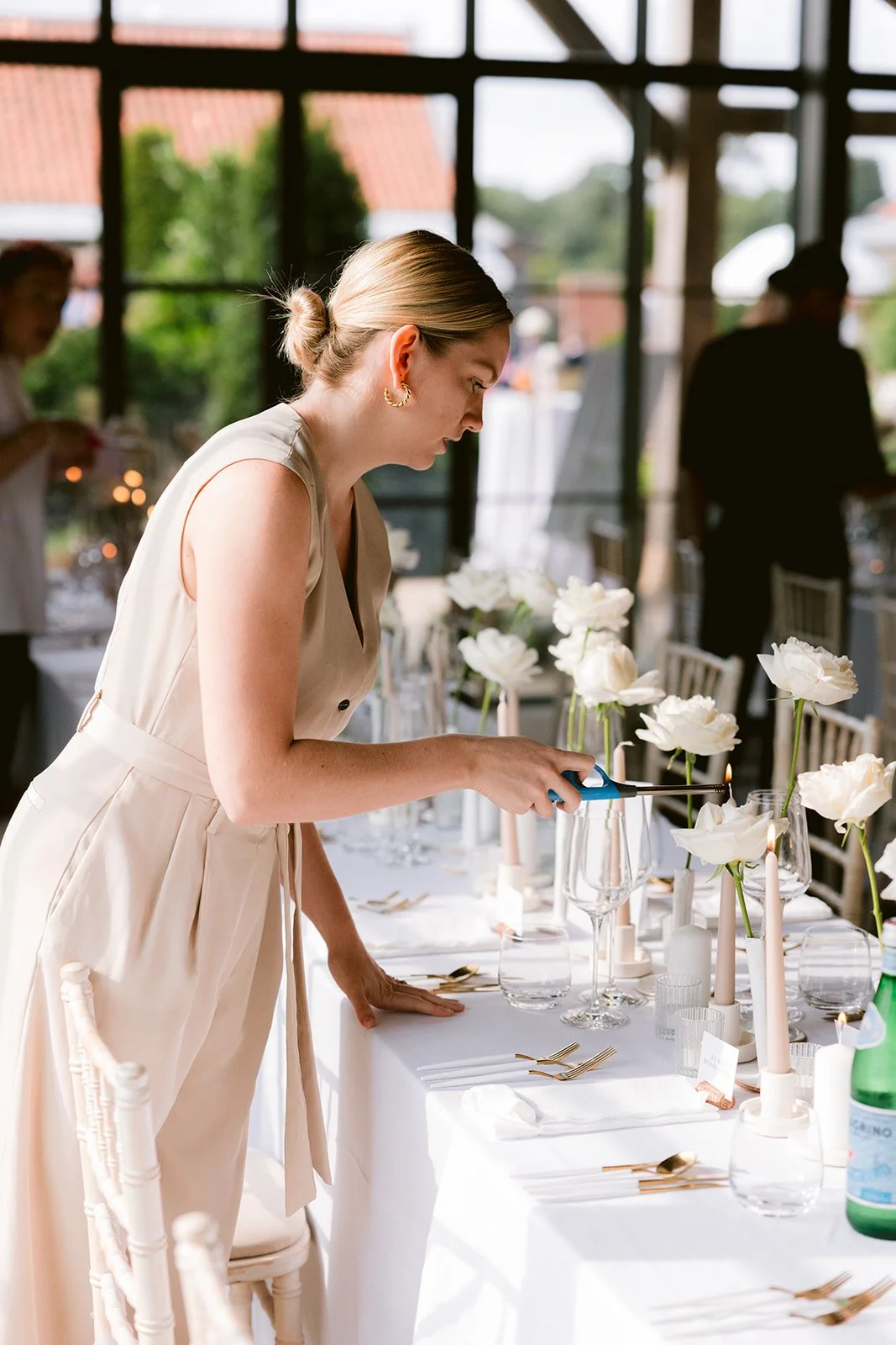 A woman with blonde hair in a bun, wearing a cream-colored dress, is lighting candles on a beautifully decorated dining table.