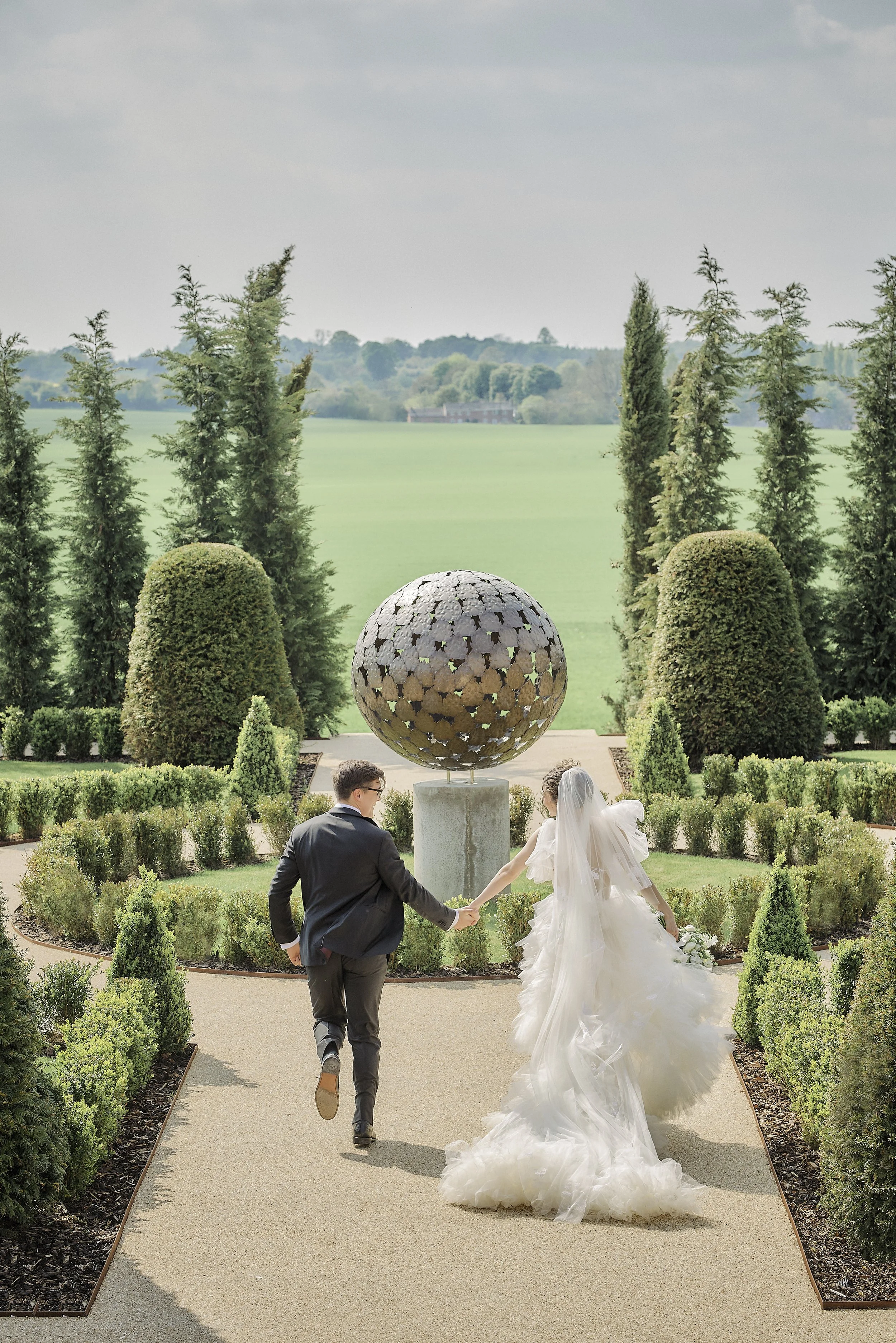 A newlywed couple holding hands and running on a garden path, with the bride wearing a white wedding dress and veil, and the groom in a dark suit, heading toward a large spherical sculpture in a formal garden with tall trees and green fields in the background.
