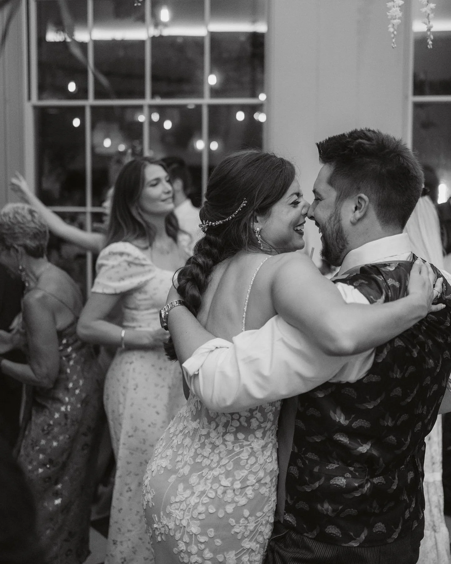 A black and white photo of a couple dancing closely at a celebration, smiling at each other, with other guests dancing and socializing in the background.