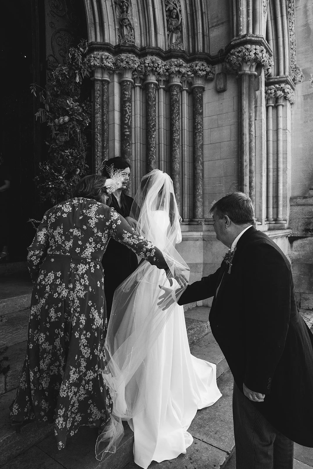 A bride in a wedding gown and veil is being assisted by three women and a man outside a church with ornate stone columns, as they prepare for the wedding ceremony.