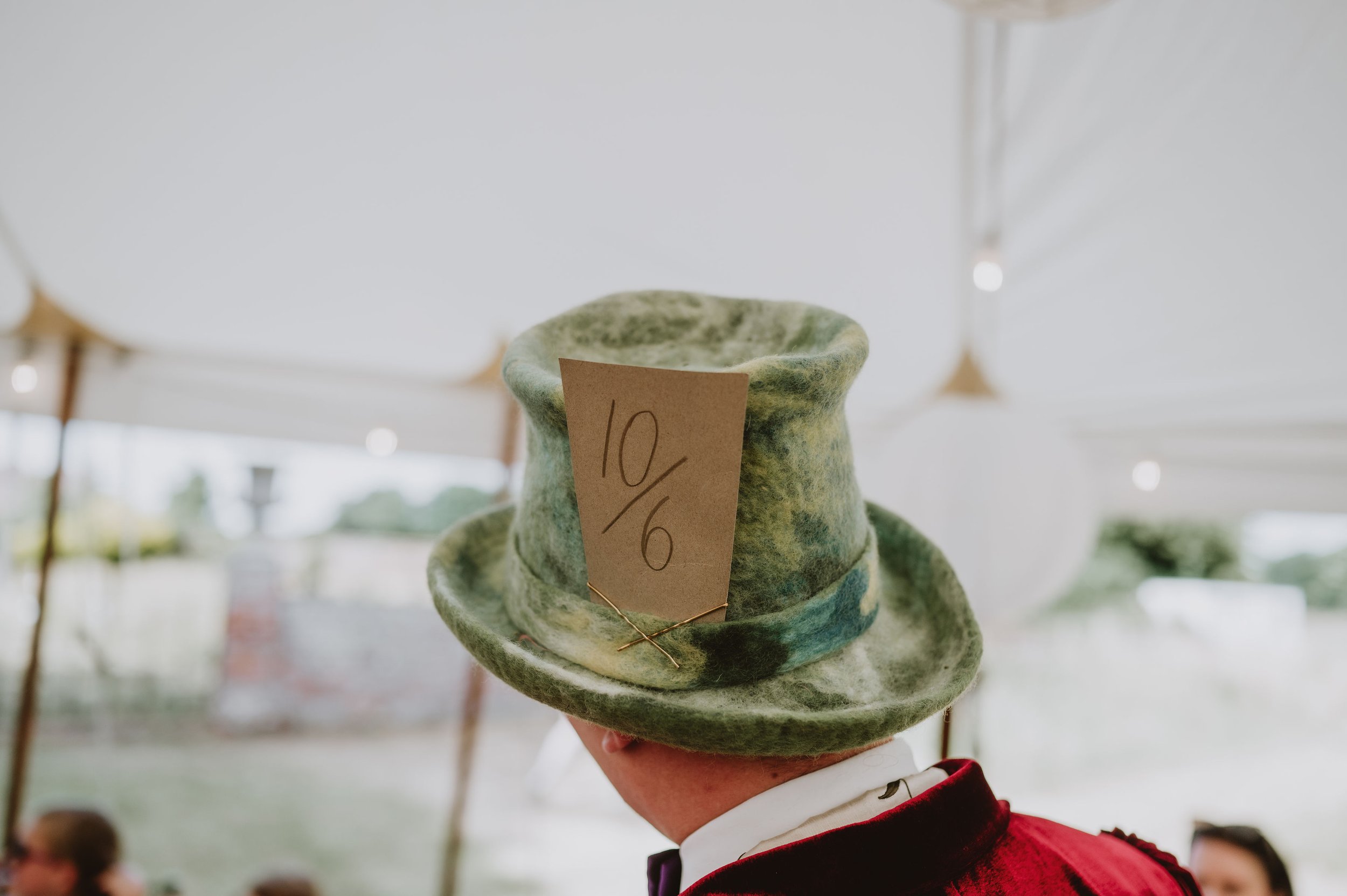 Person wearing a green felt top hat with a handwritten note indicating 10/6 pinned to it during a daytime outdoor event.