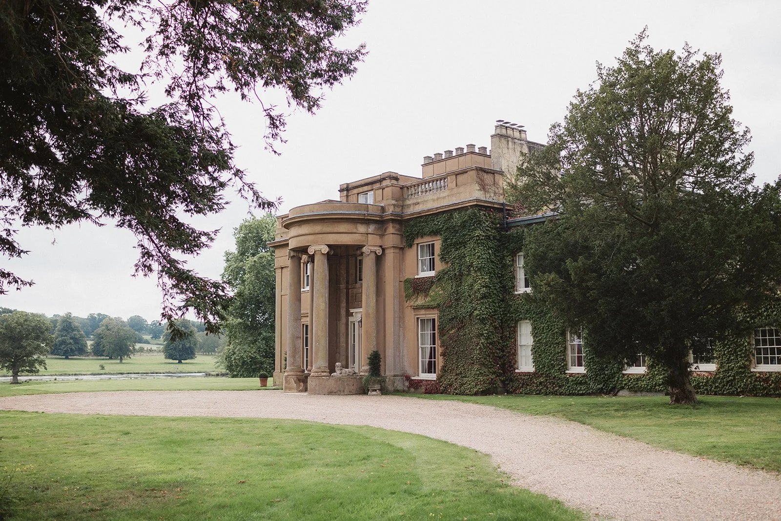 A large historic mansion with classical architecture, surrounded by green lawn and trees, with an overcast sky.